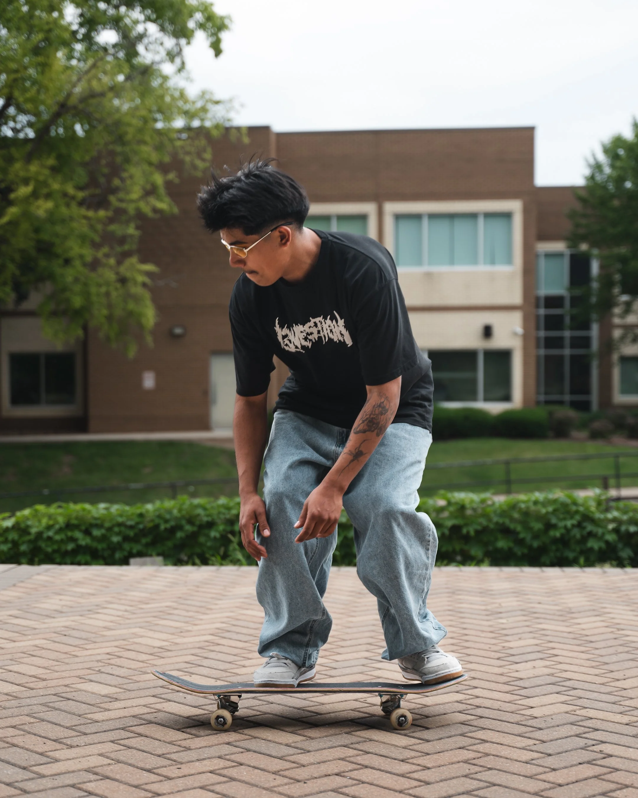 A young man with black hair, wearing yellow-tinted sunglasses and a black graphic t-shirt, is skateboarding outdoors on a paved area in front of a residential building with trees and bushes around him.