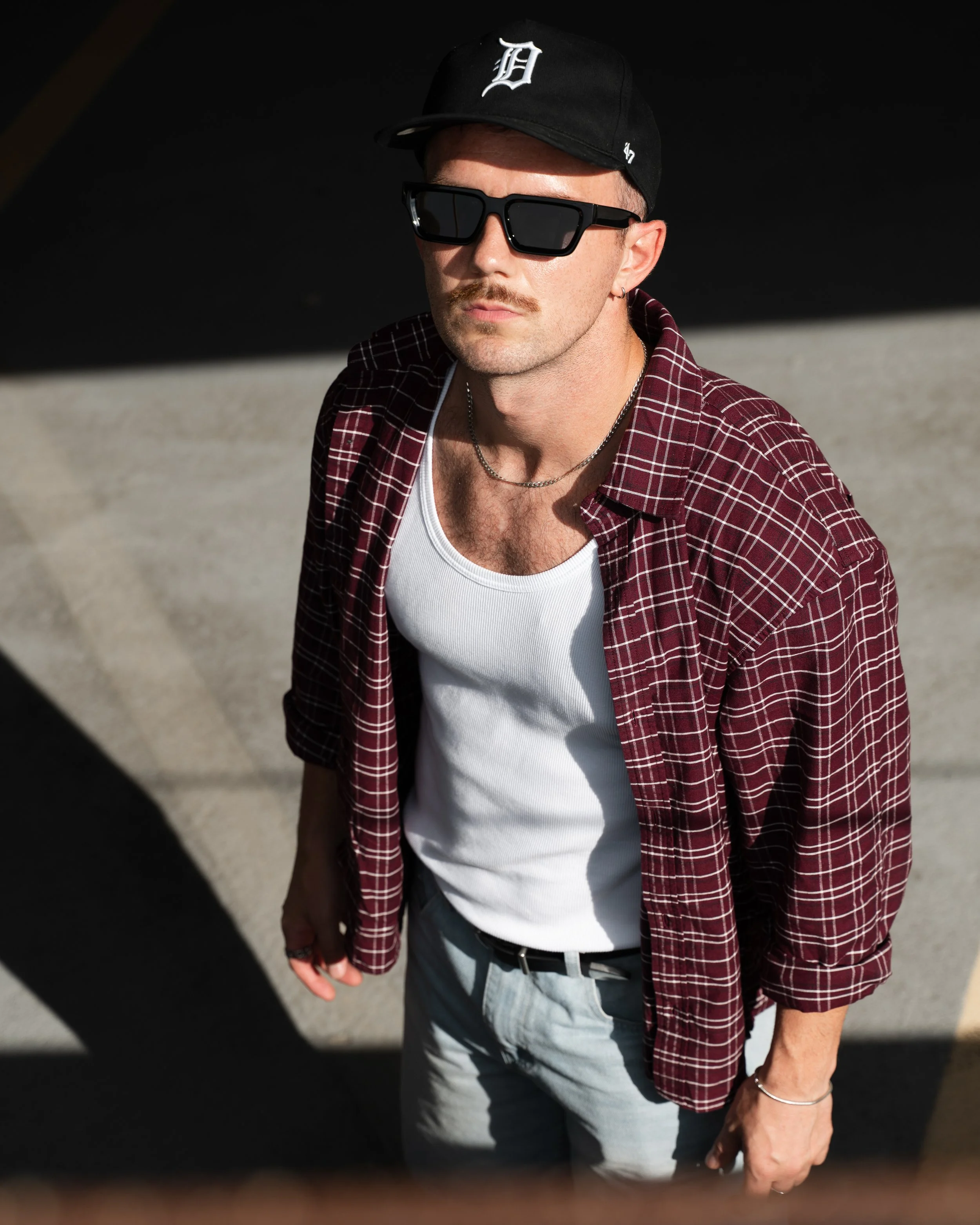 A person wearing a black Detroit Tigers cap, black sunglasses, a red plaid shirt over a white tank top, gray pants, and accessories including a silver chain and bracelet, standing outdoors with a shadow cast on their face.