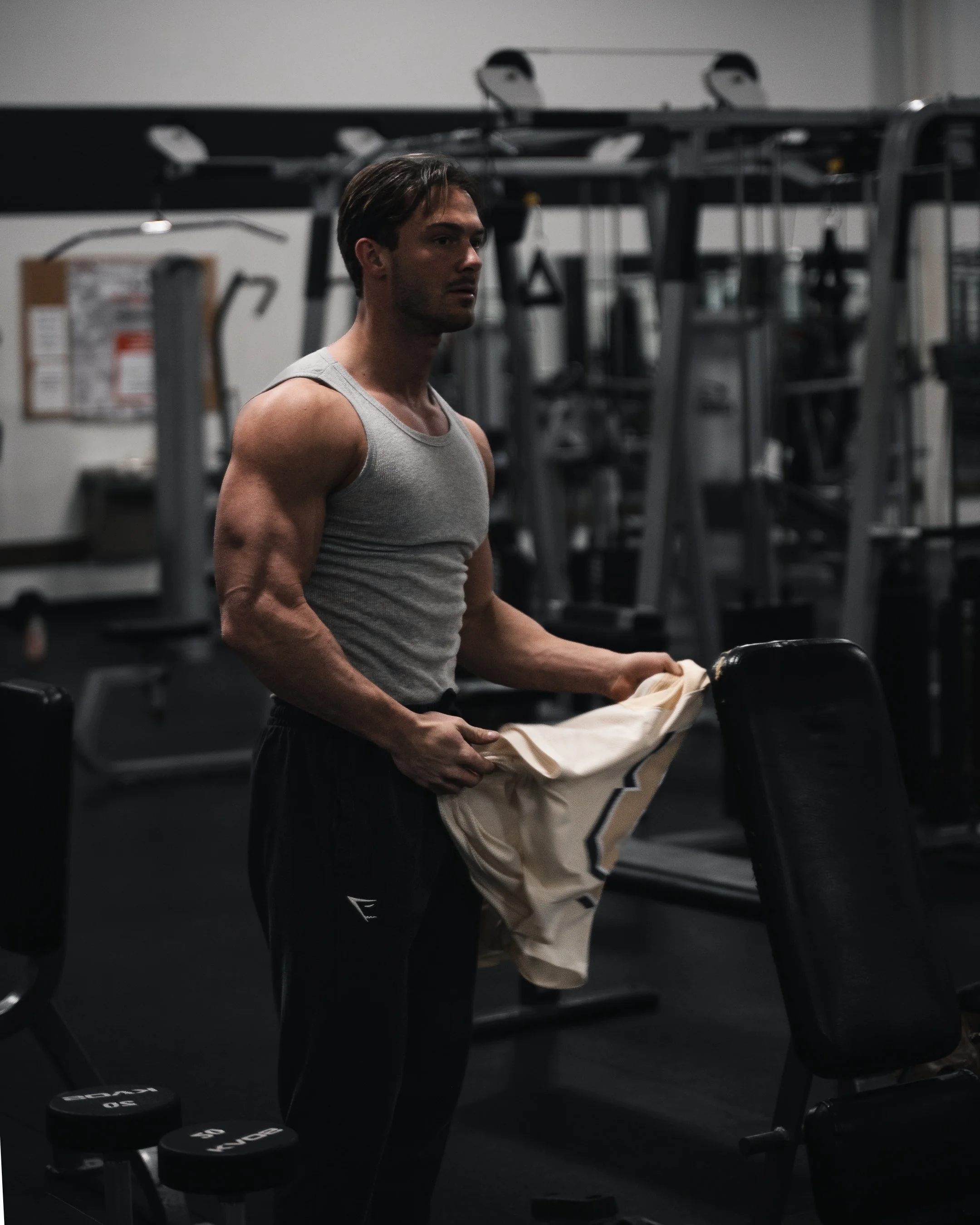 A man in a gray tank top holding a beige towel in a gym surrounded by exercise equipment and dumbbells.