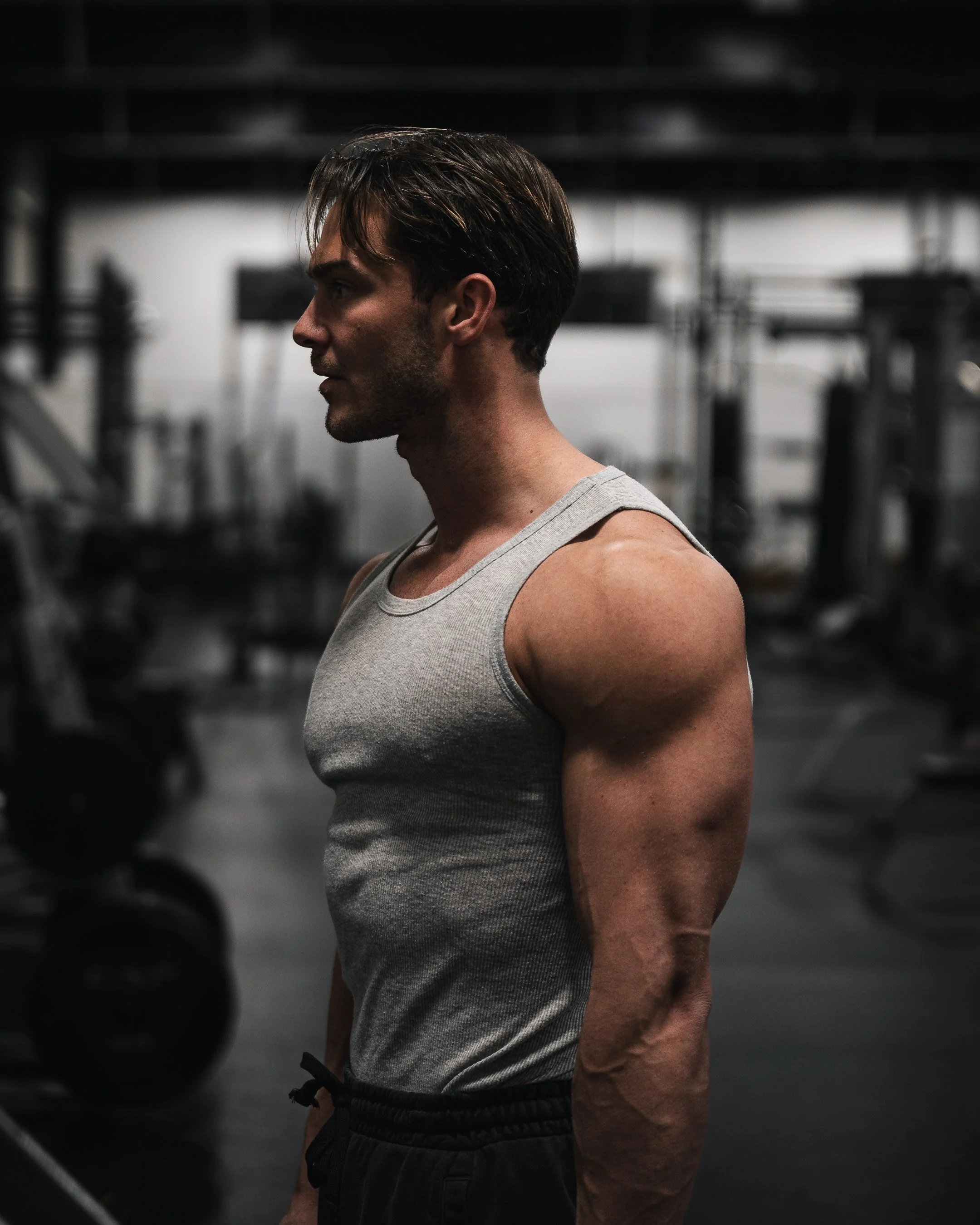 A side profile of a muscular man wearing a gray tank top and black shorts in a gym, with blurred gym equipment in the background.