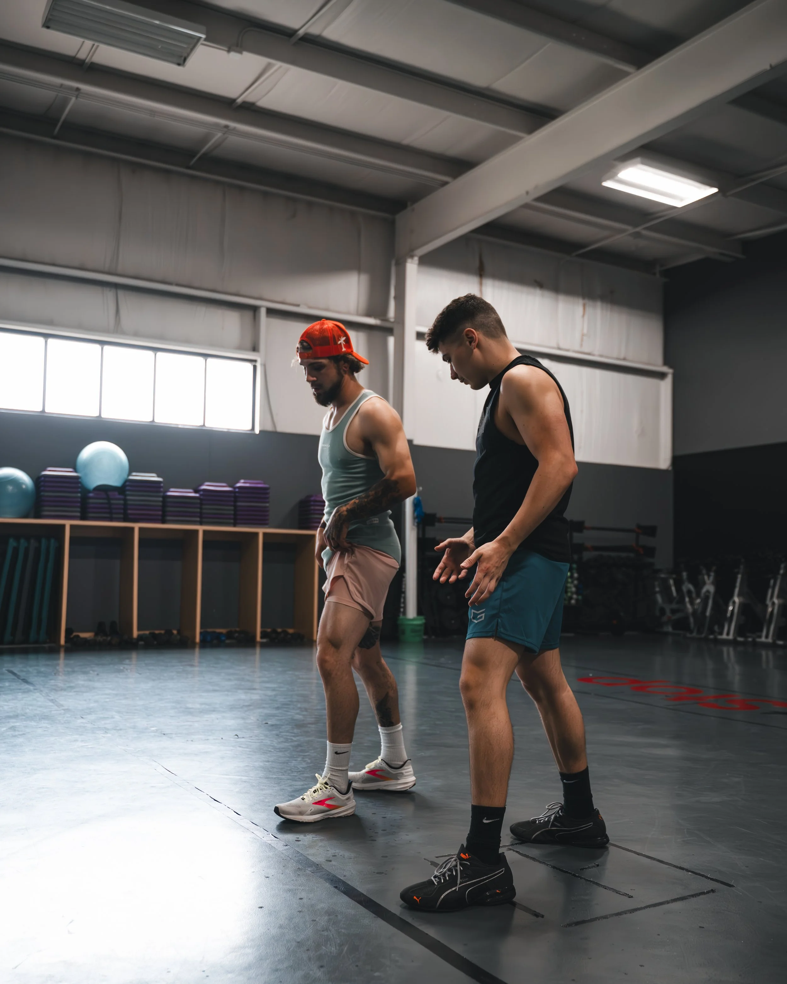 Two young men standing in a gym or fitness studio, looking downward and possibly preparing for exercise. They are dressed in athletic clothing, and their shoes are prominent with one wearing white and the other black sneakers. The gym has a gray floo