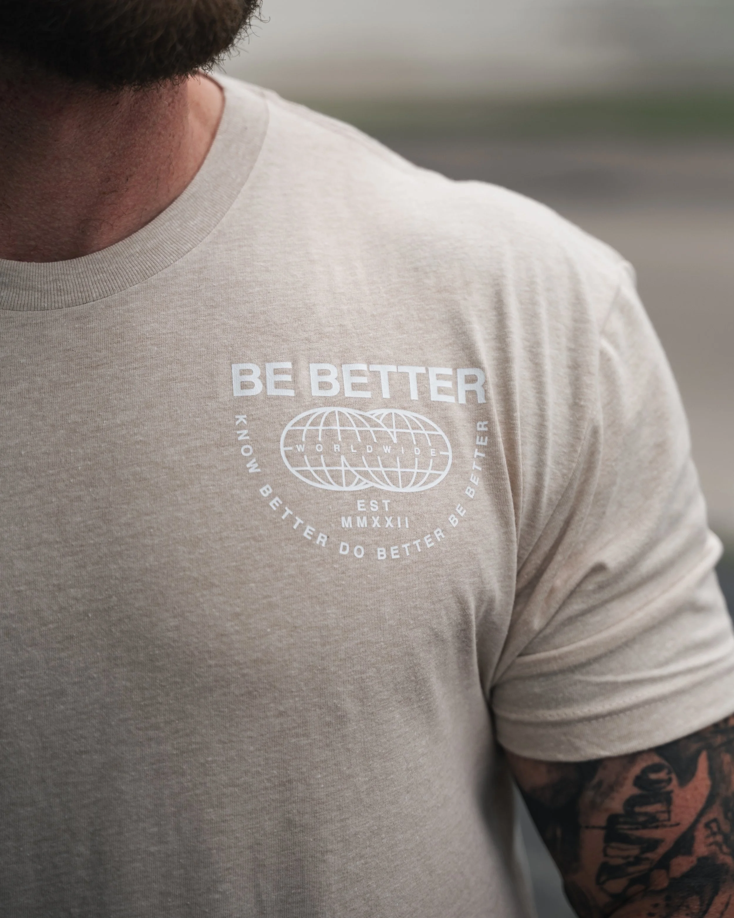 Close-up of a person wearing a beige T-shirt with white text and a globe graphic. The person's shoulder and tattooed arm are visible, with dark hair and beard partially seen.