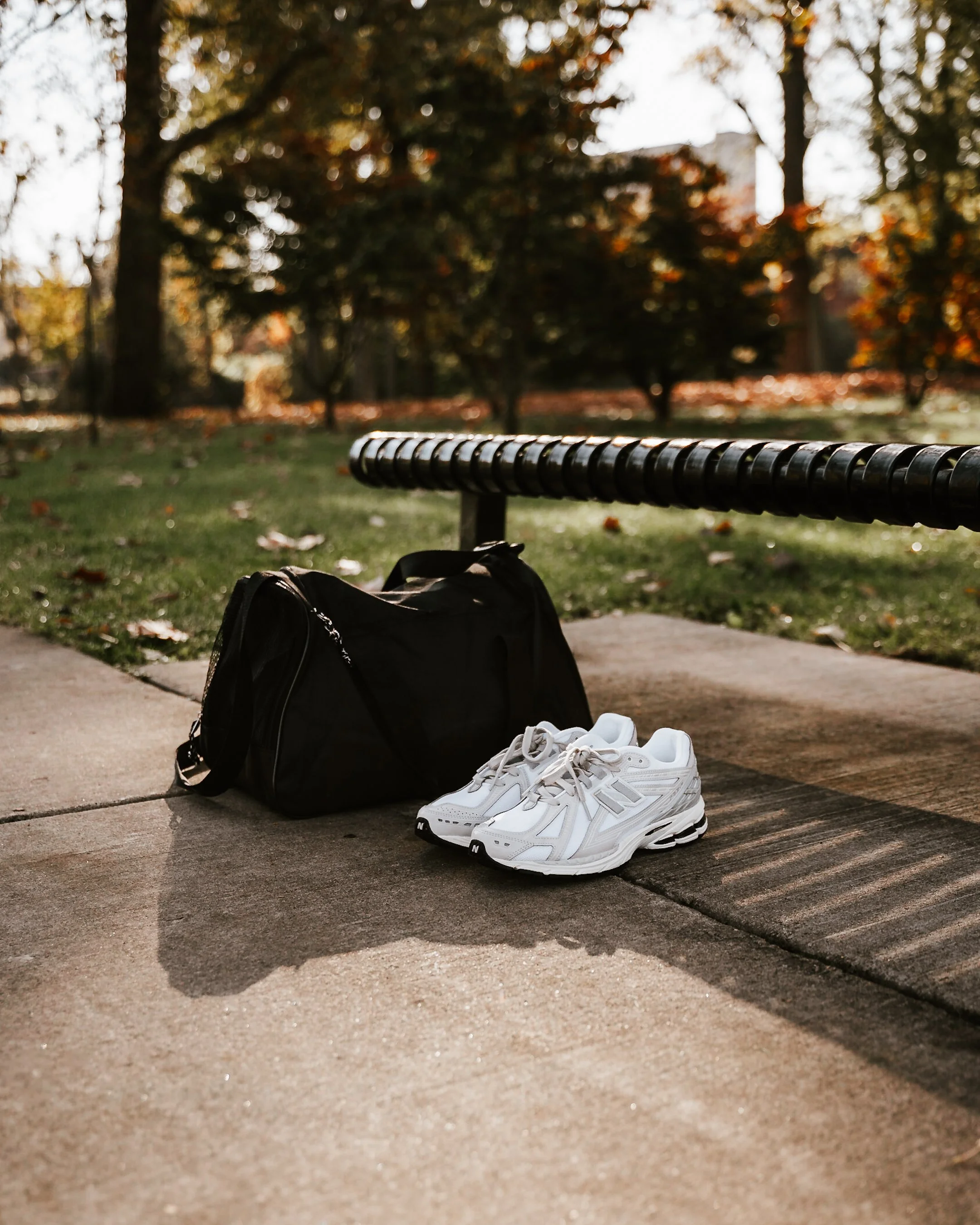 White sneakers, a black backpack, and a cap on the ground in front of a park bench in a park with trees and fallen leaves