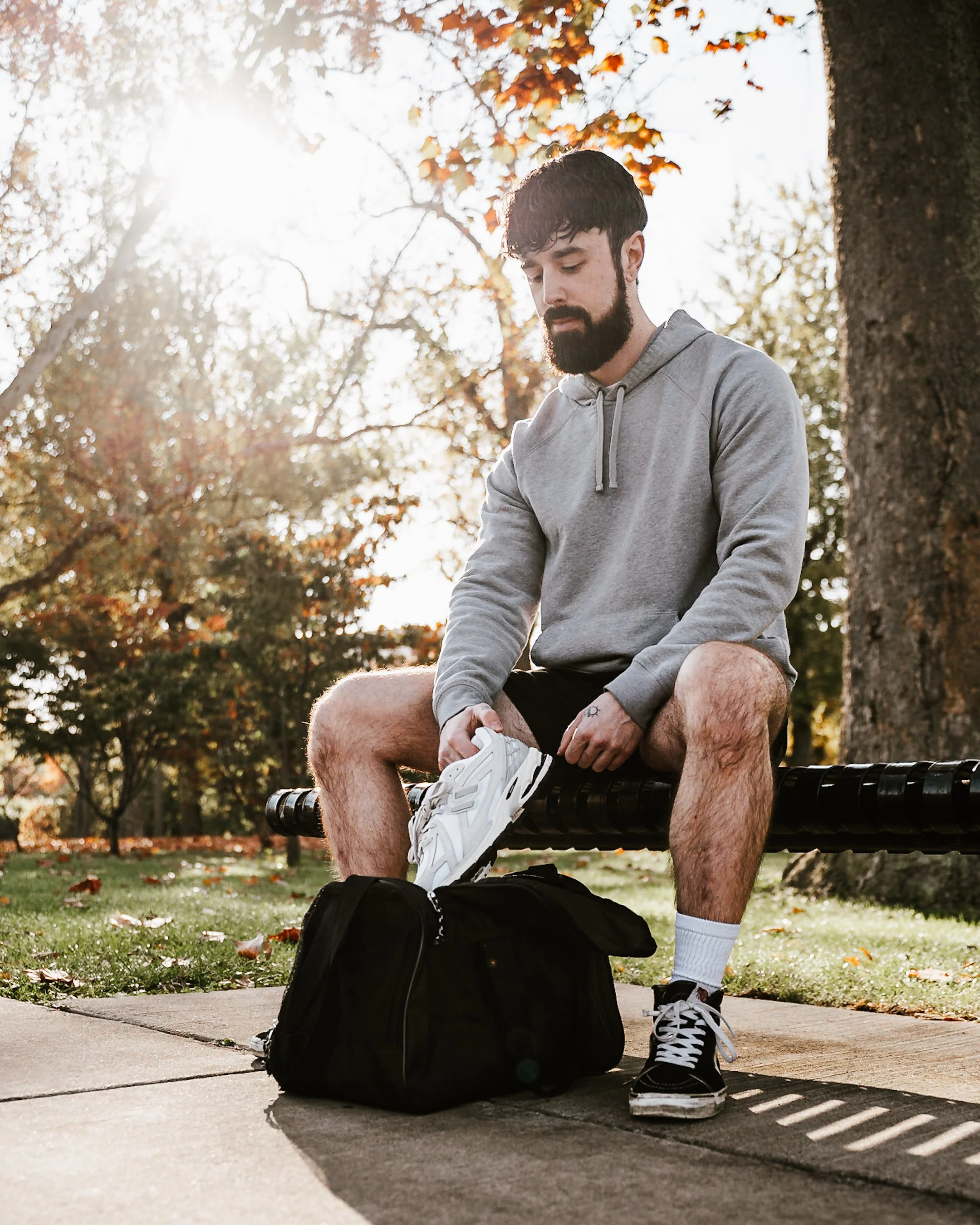 A young man with dark, curly hair and a beard sitting on a park bench, adjusting his white sneaker while looking down. He is wearing a light gray hoodie, black shorts, white socks, and black and white sneakers. A black backpack is on the ground in fr