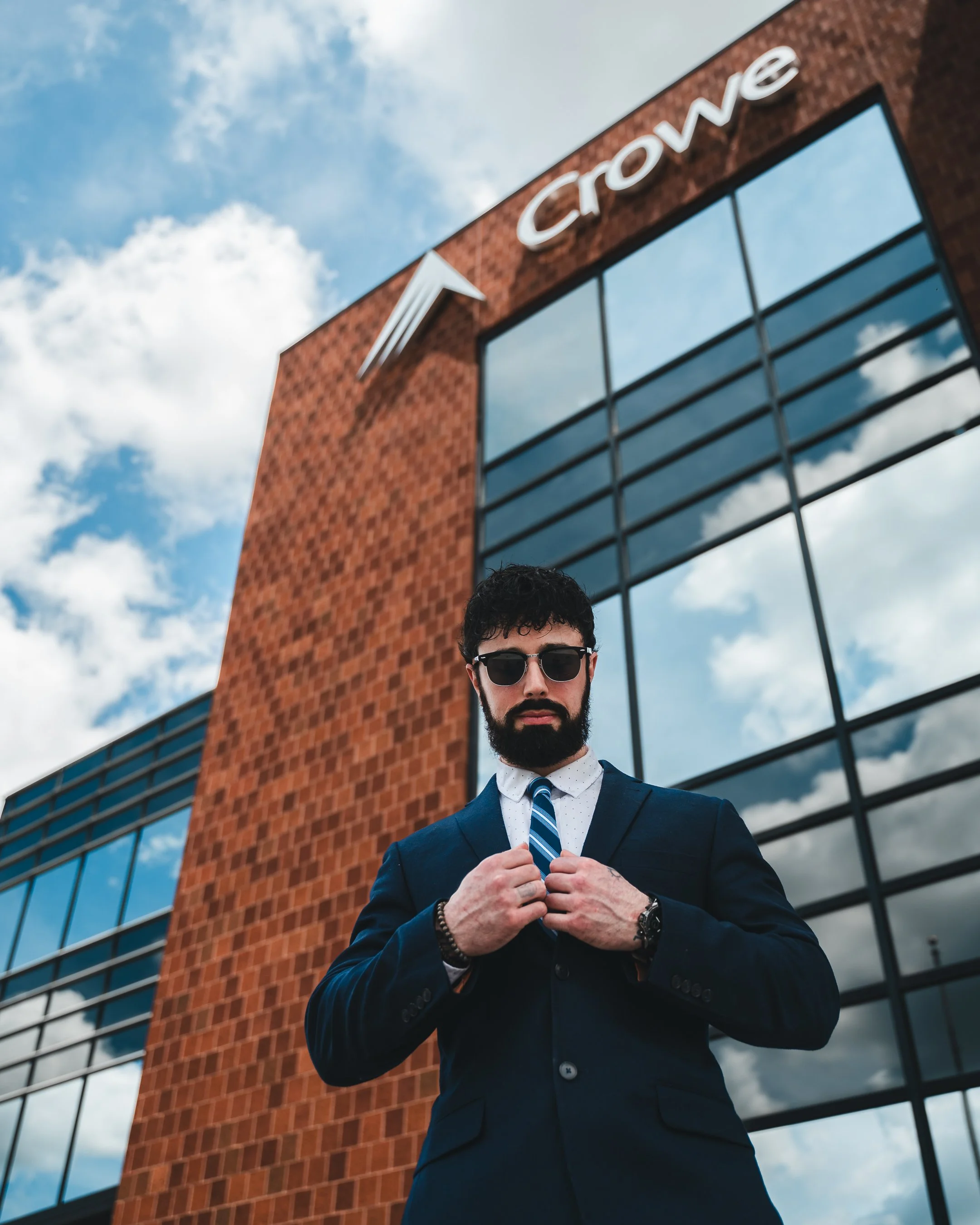 A man with a beard and sunglasses, wearing a suit and tie, is adjusting his suit jacket in front of a modern building with large glass windows and a brick facade, with the word 'Crown' visible on the building.