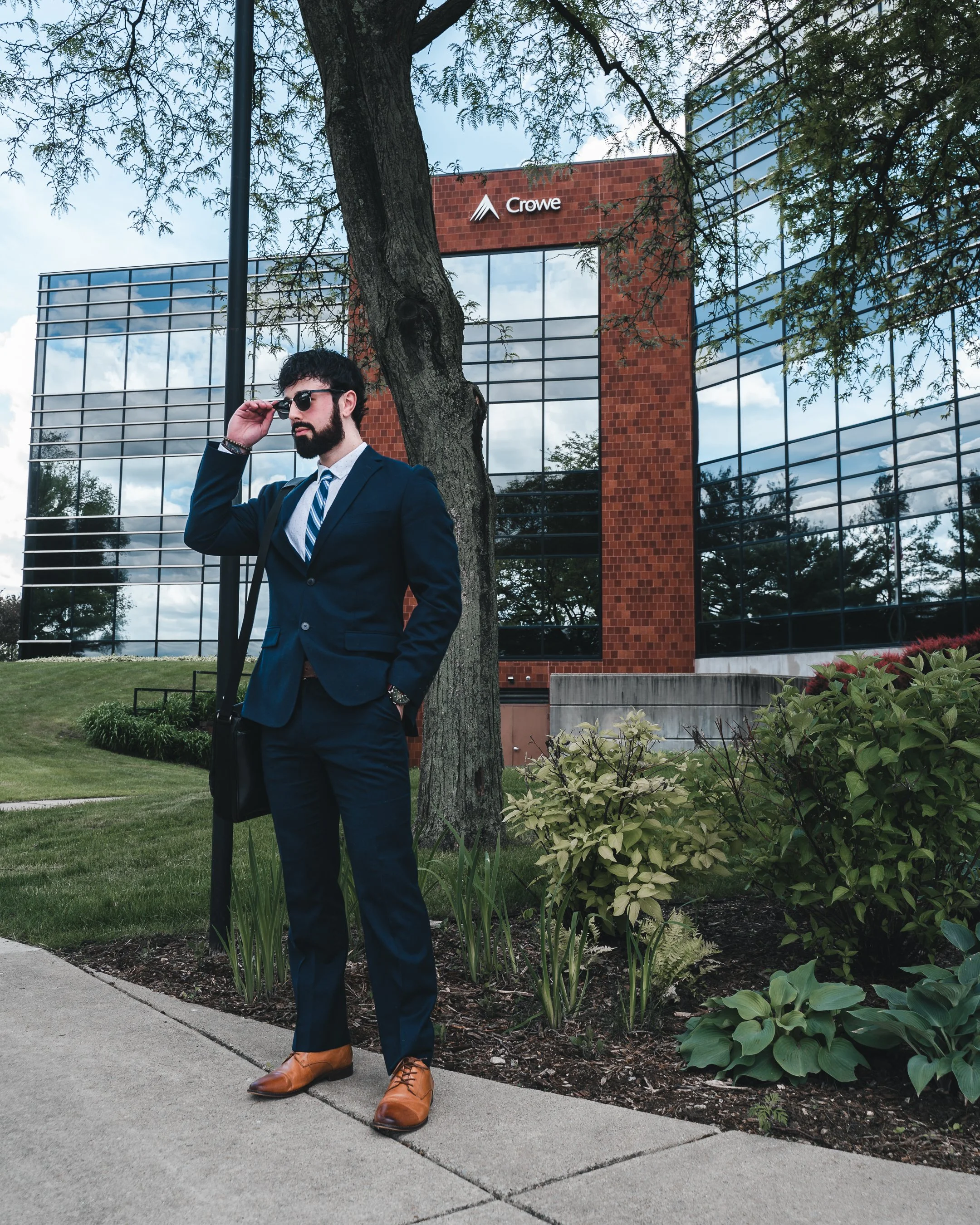 A man in a navy suit and brown shoes standing next to a tree and bushes outside a modern glass office building with the Crowe logo.