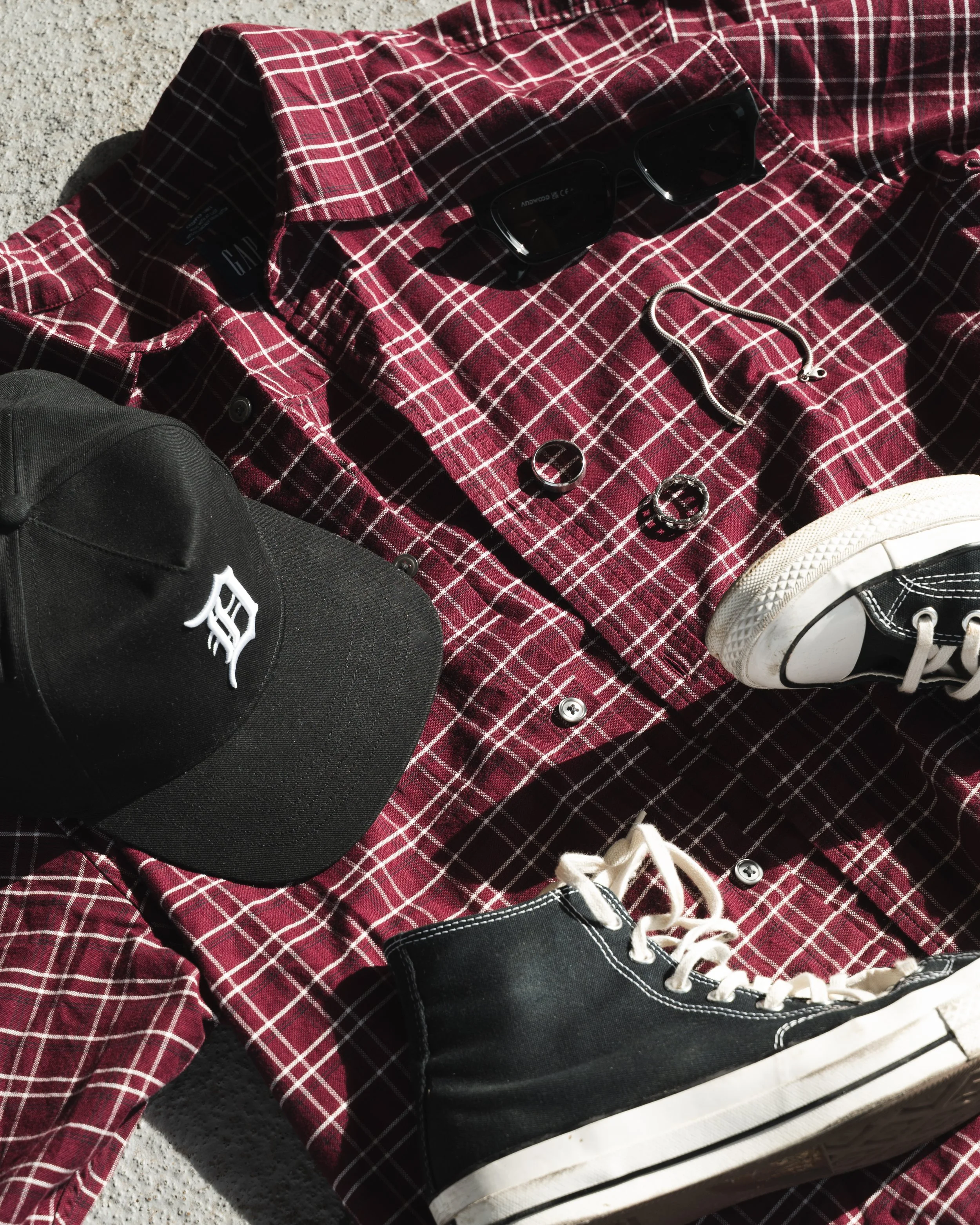 A red plaid button-up shirt, black sneakers with white soles, black sunglasses, a silver chain, and two rings are laid out on sand.