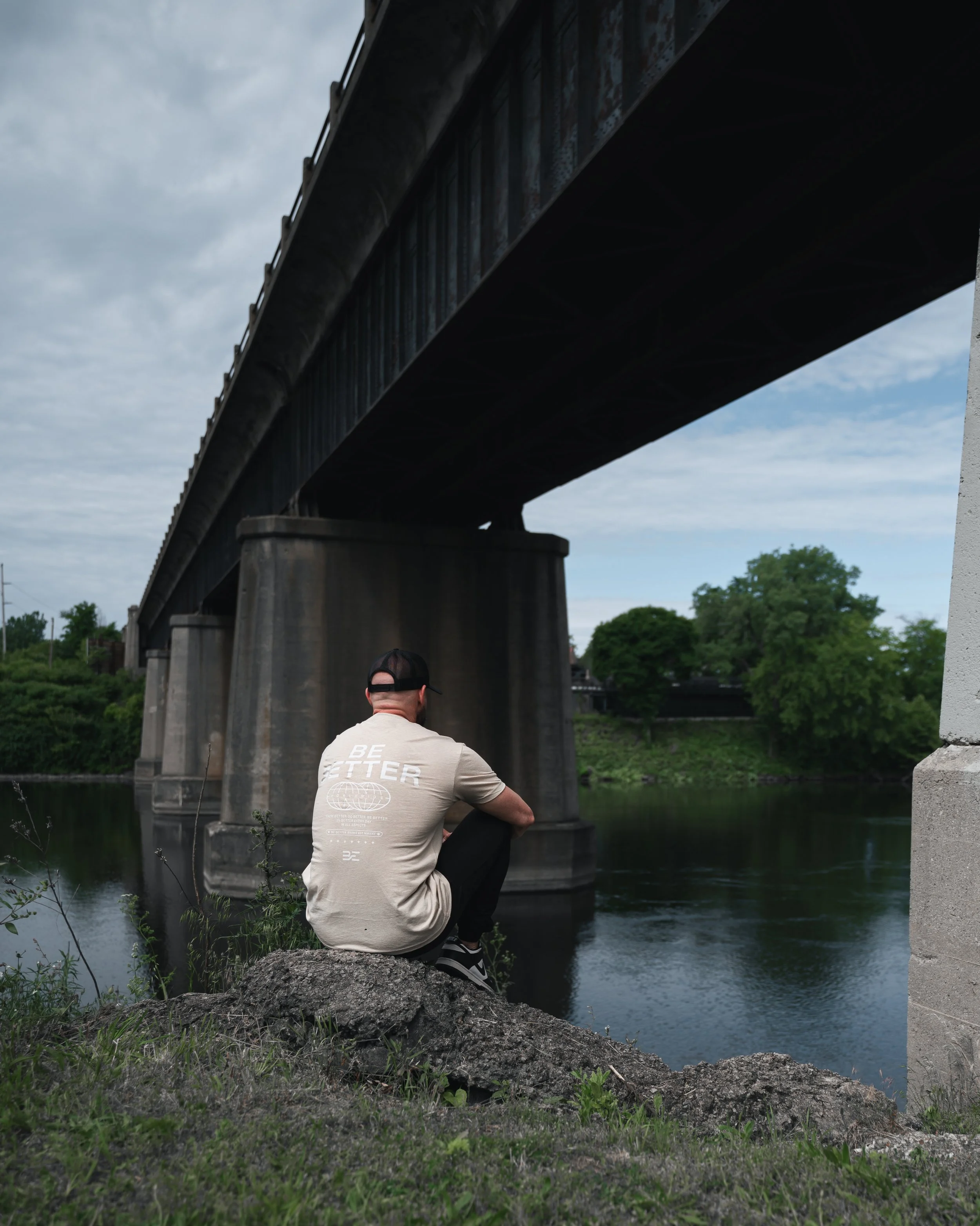 A man sitting on a rock by a river, looking at a bridge overhead, with trees in the background and cloudy sky.