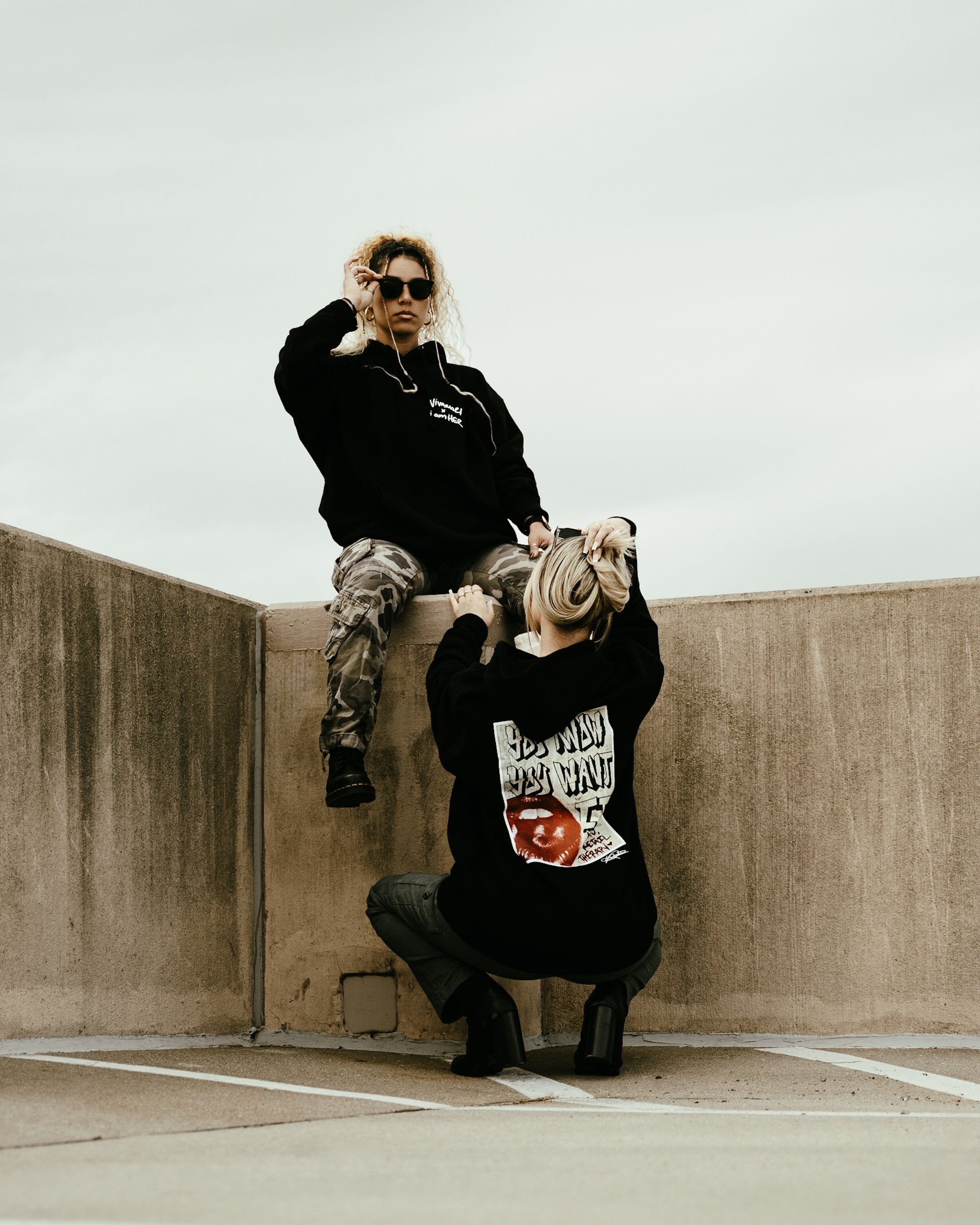 A woman with curly hair, wearing sunglasses and a black sweatshirt, sitting on a concrete ledge, holding a leash and petting a dog below her. Another person with blonde hair, wearing a black hoodie, is crouched beside her, reaching up to the woman.