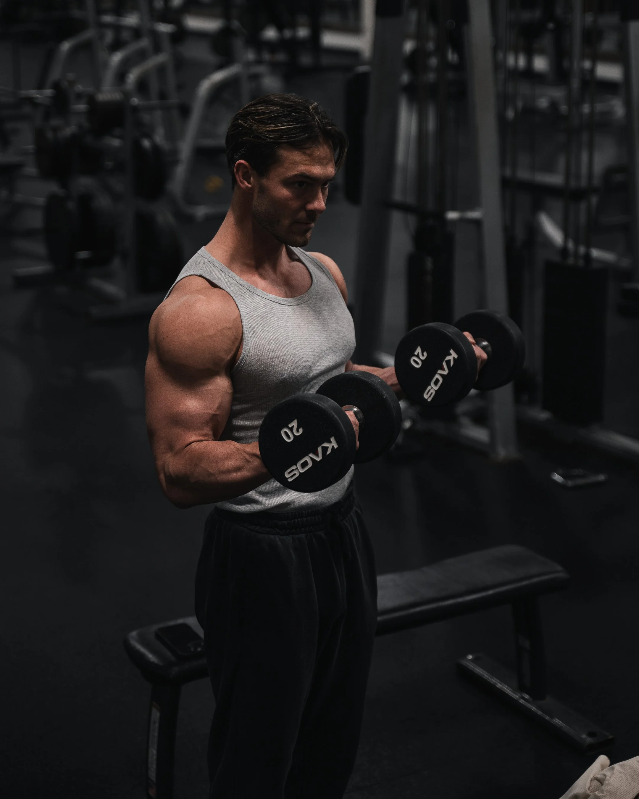 A man lifting black dumbbells in a dark gym, wearing a gray tank top and black pants.