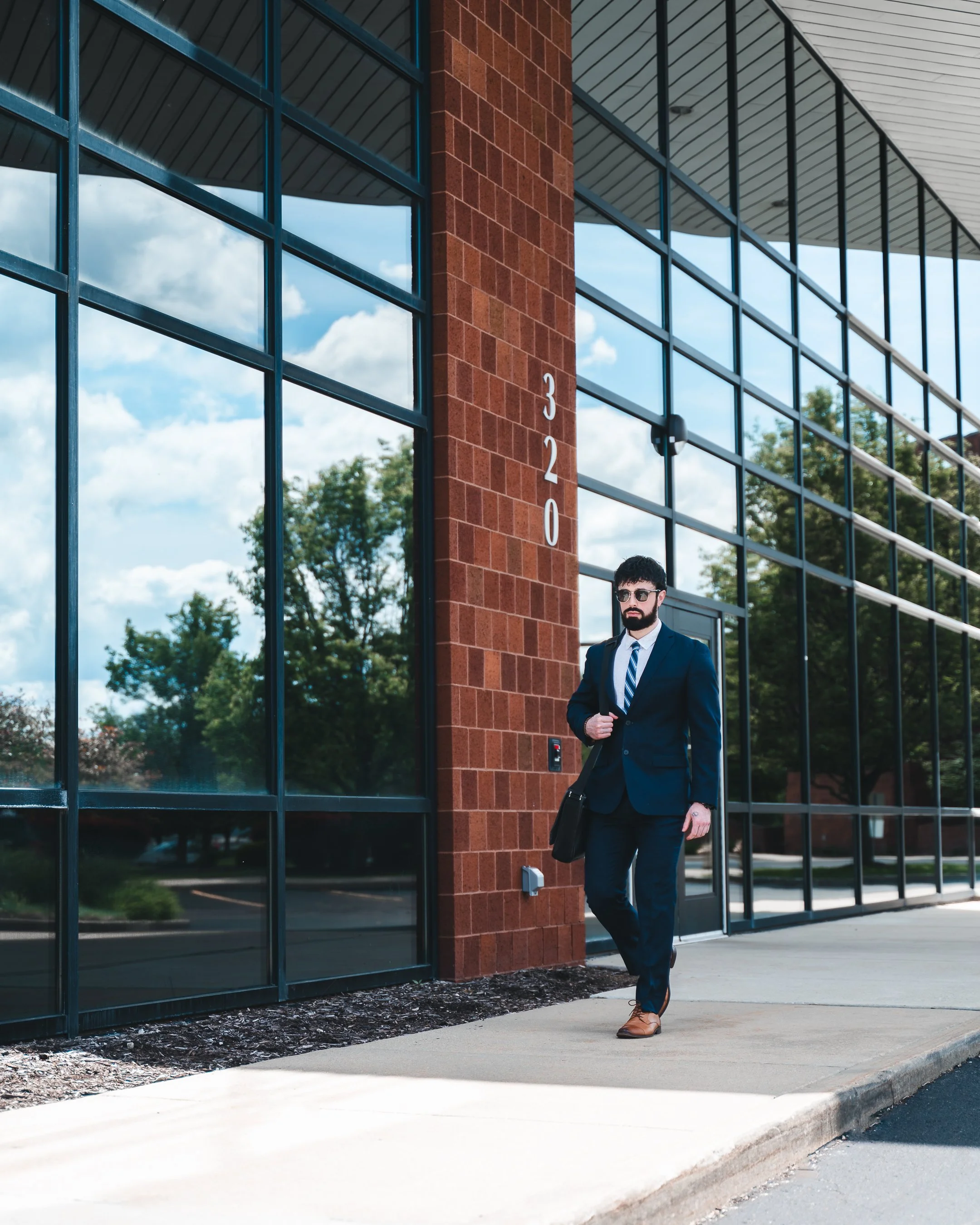 A man in a suit with sunglasses and a briefcase walking outside a modern building with large glass windows and a brick column labeled 320.