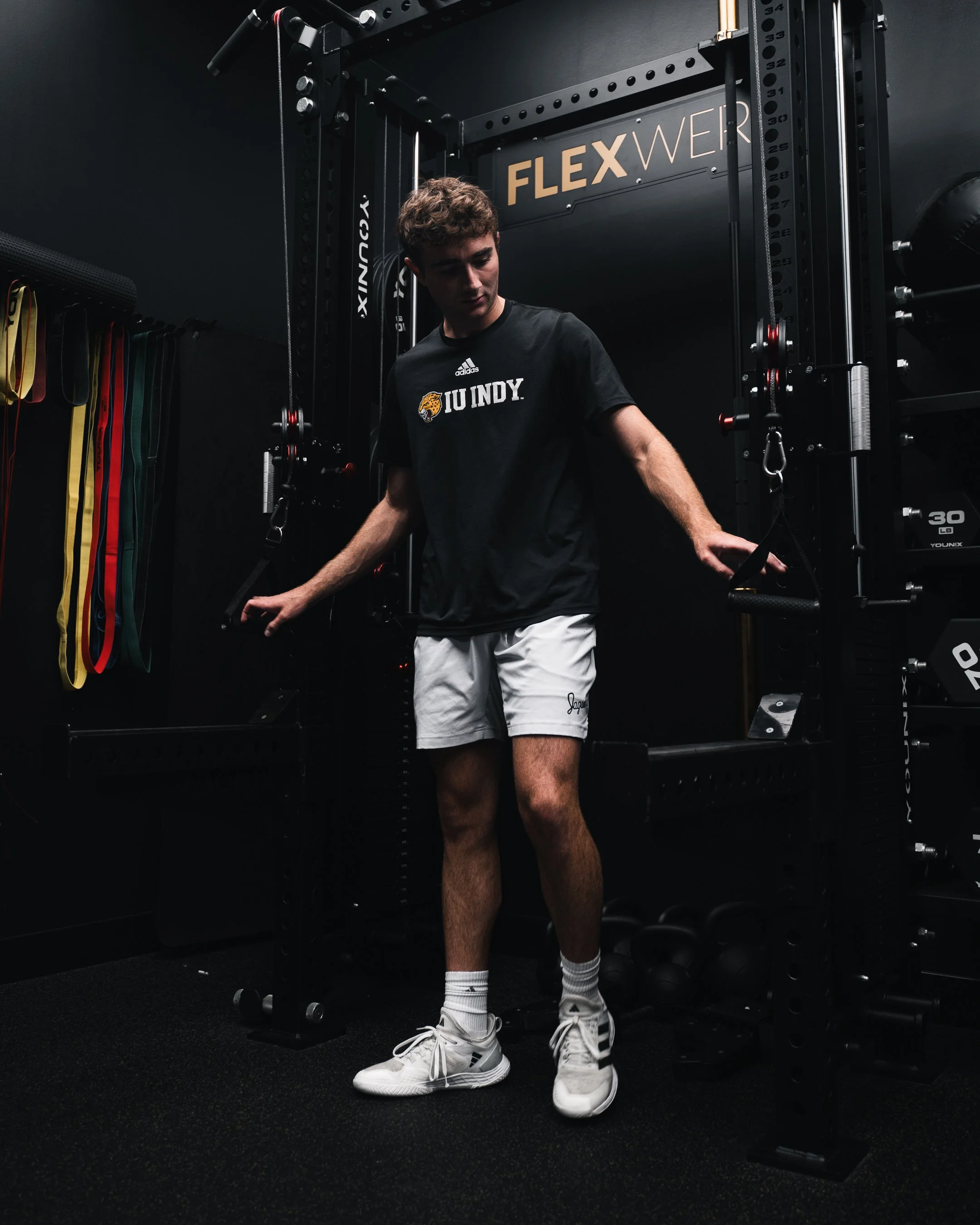 A young man stands in a gym next to strength training equipment, wearing a black t-shirt with 'IU INDY' and a tiger logo, white shorts, and white sneakers with black stripes.