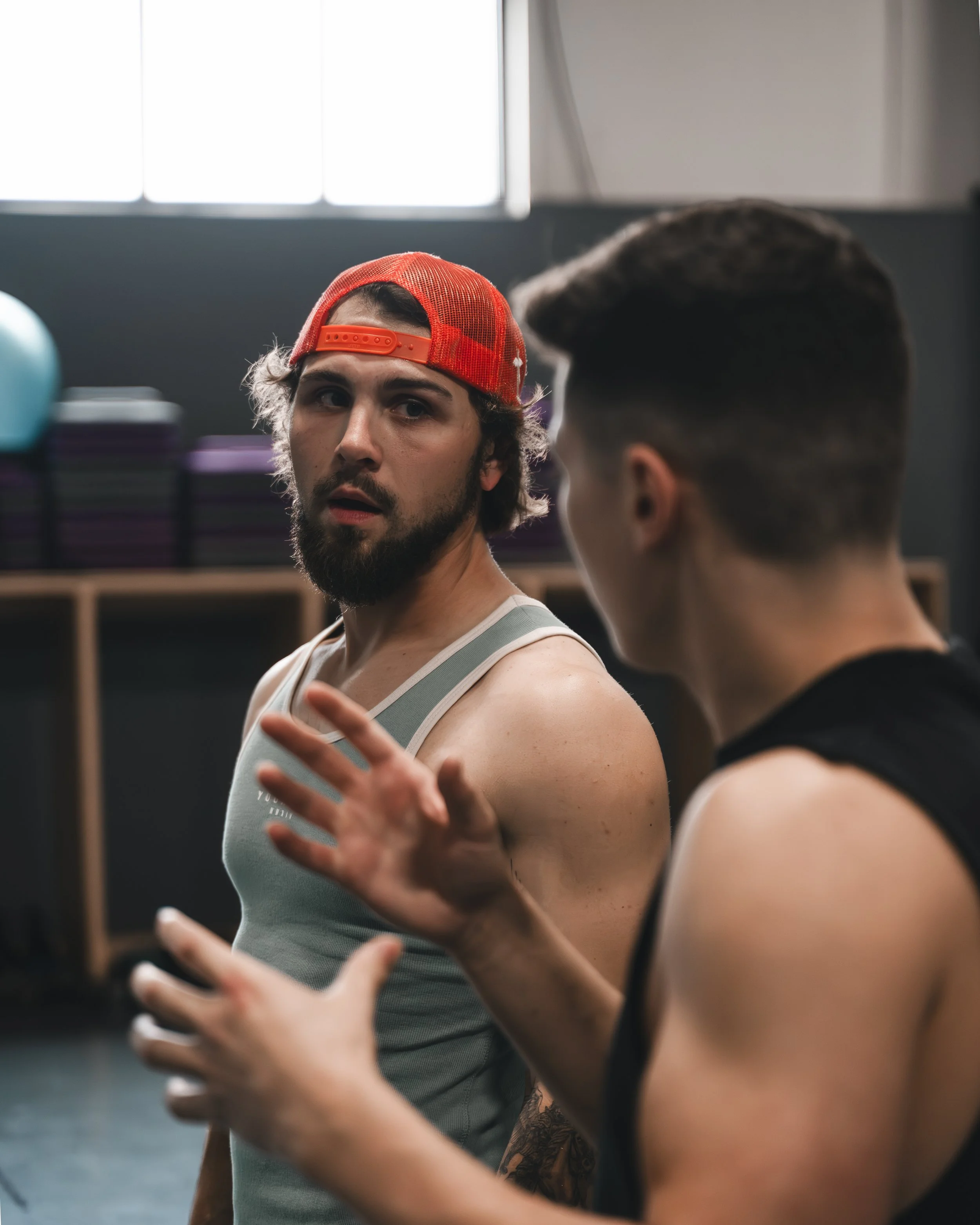 Two men having a conversation in a gym. One man wearing a red mesh cap backward and a gray tank top, the other man with dark hair and a sleeveless black shirt.