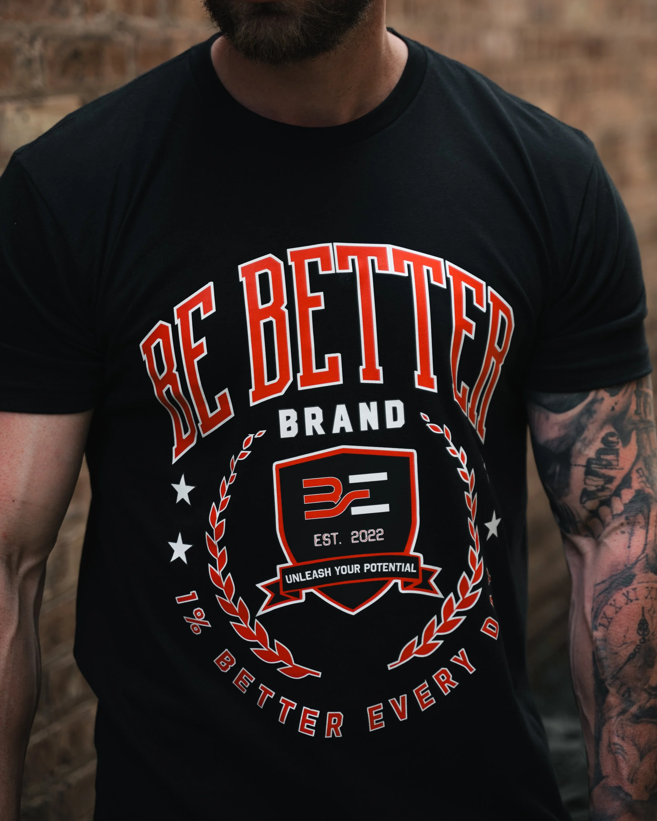 Close-up of a man wearing a black T-shirt with a large red and white graphic design and text reading 'Authentic', 'Brand', 'EST. 2022', and 'Unleash Your Potential.' The man has a beard and tattoos on his arms, and the background features a brick wal