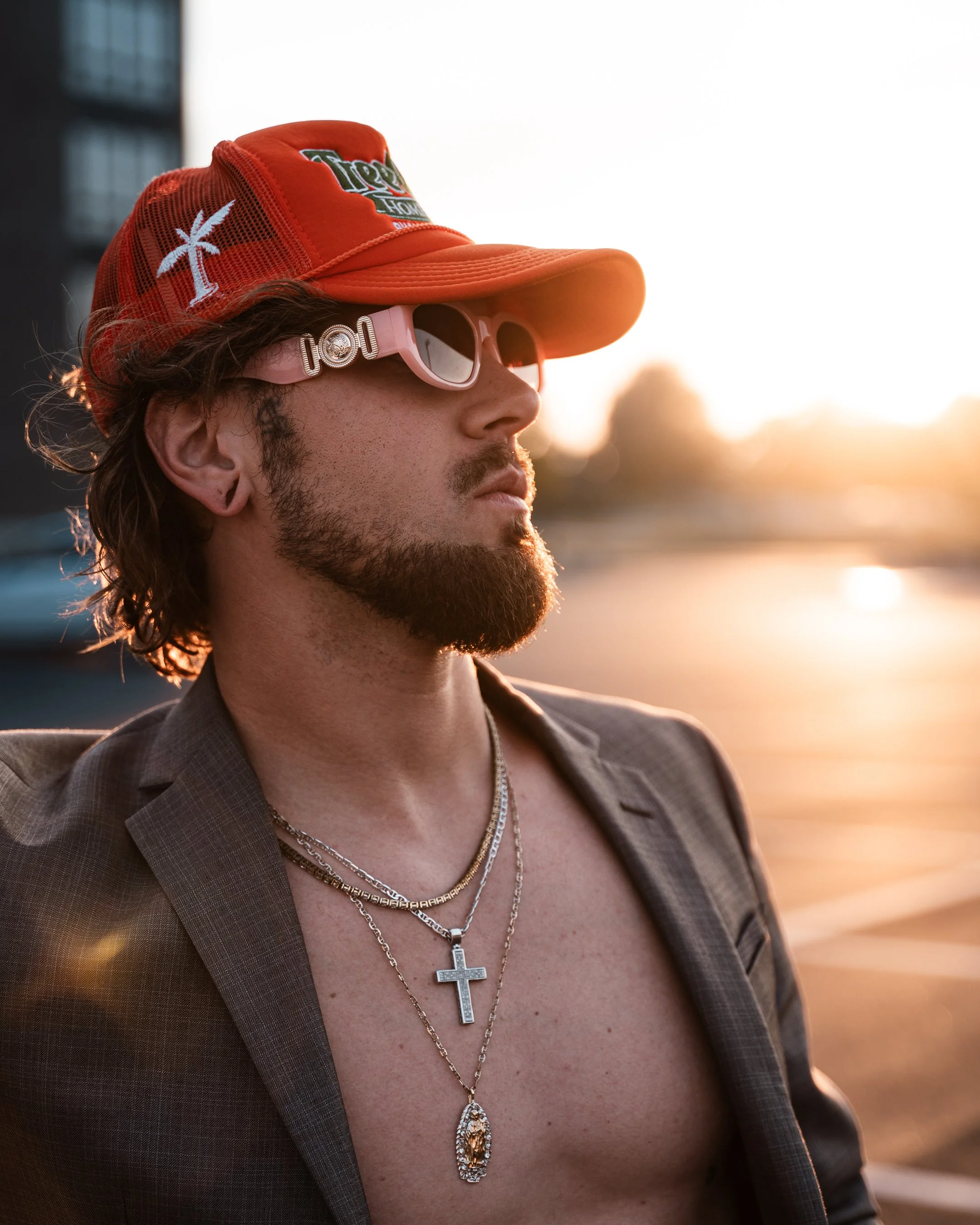 A man with long dark hair, mustache, and beard, wearing a brown plaid jacket, layered gold necklaces, and pink sunglasses, is seen in profile wearing an orange baseball cap with a palm tree and logo, during sunset outdoors.