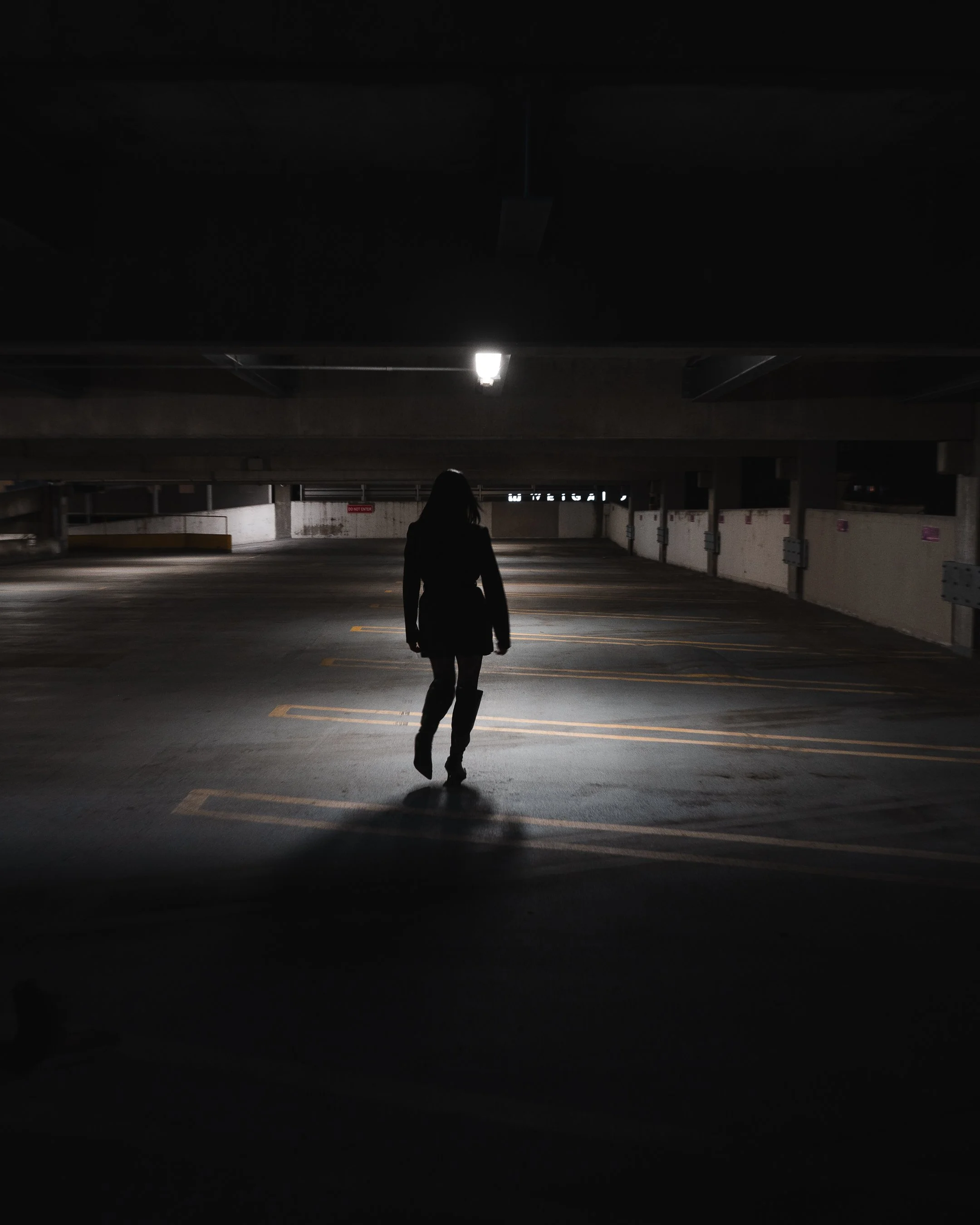 A person is walking in an underground parking garage illuminated by a ceiling light, with their back facing the camera.