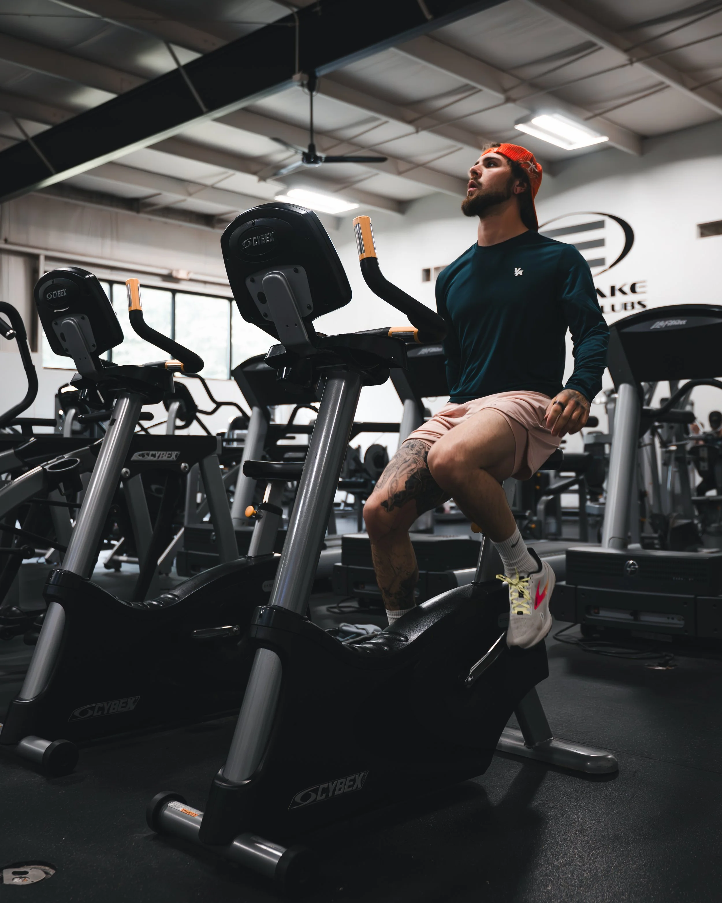A man with tattoos, wearing a red cap backwards, a dark sports shirt, beige shorts, and white sneakers with colorful accents, is sitting on a black exercise bike in a gym, looking up. There are multiple exercise bikes and gym equipment in the backgro