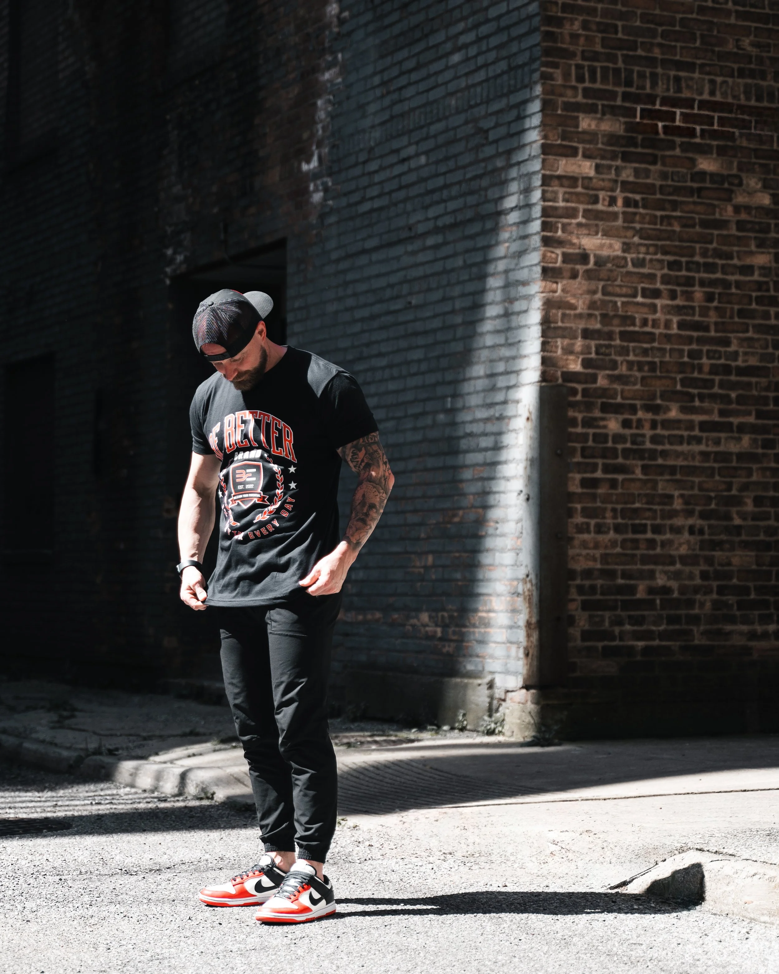A man wearing a grey cap, glasses, a black t-shirt, and black pants is looking down while standing outdoors on a street in front of a brick wall. He is wearing red, white, and black Nike sneakers.