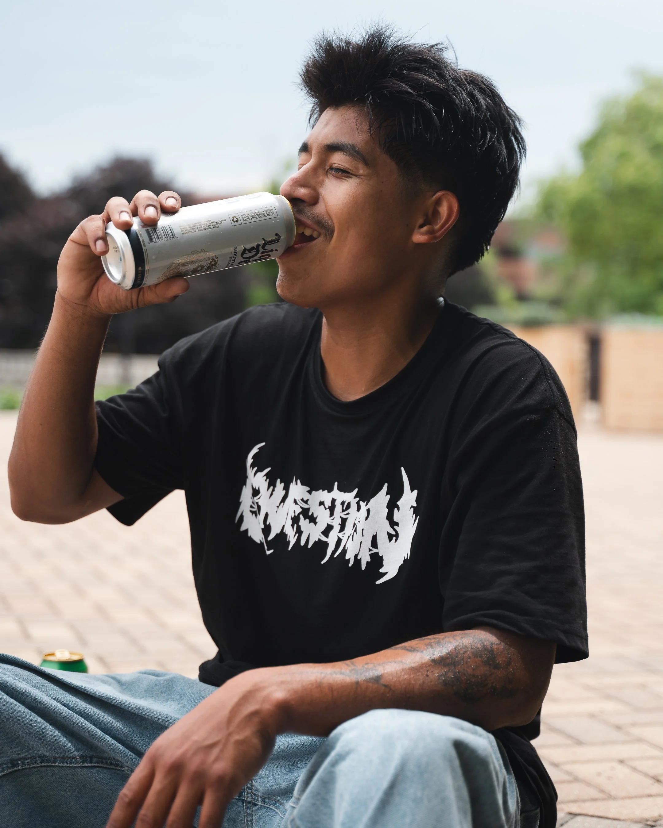 A young man with dark hair and a tattoo on his arm, wearing a black graphic T-shirt, sitting outdoors and drinking from a silver can of soda or beer. The background includes blurred trees and a brick building.