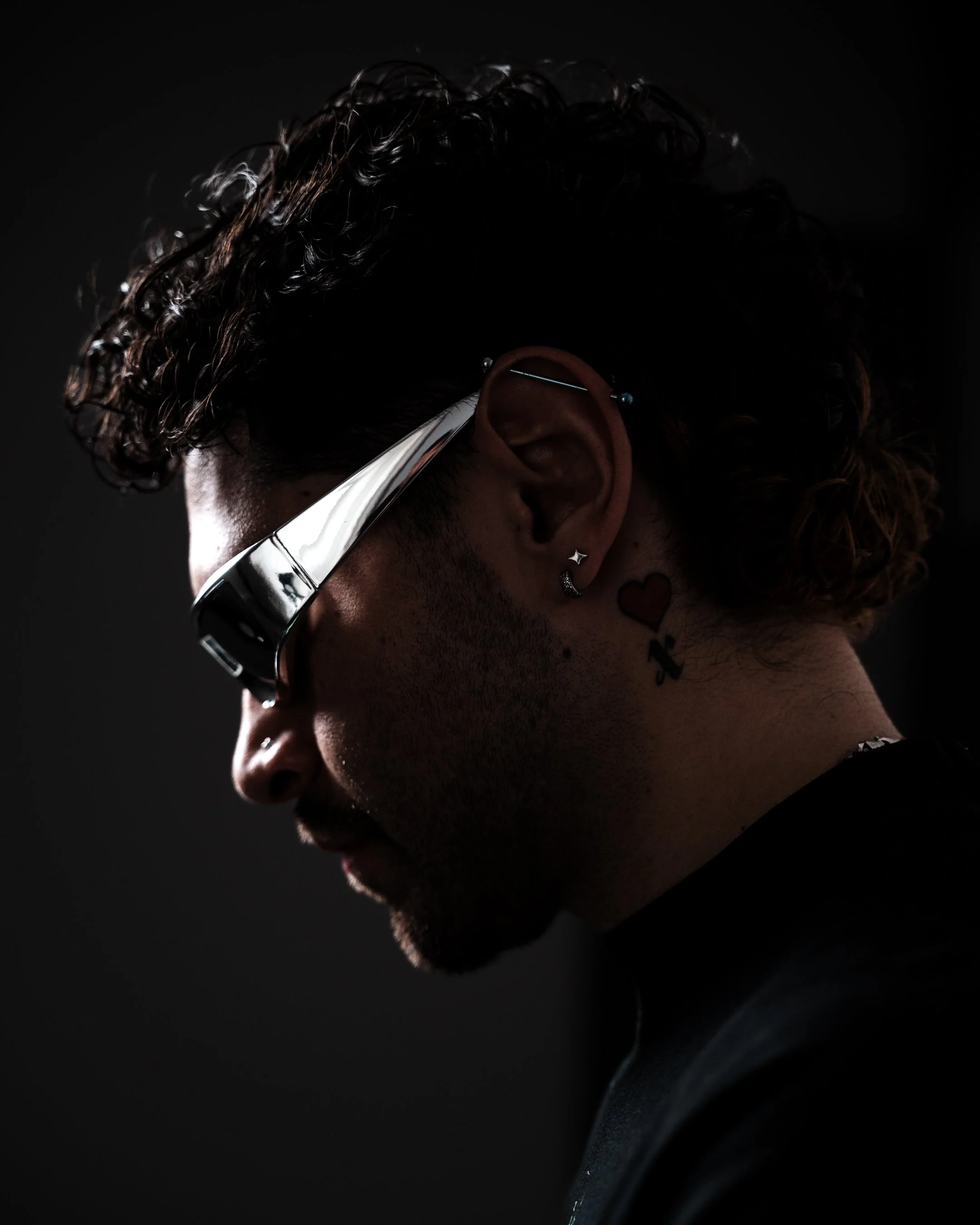 A person with curly hair is wearing large glasses and multiple earrings, including a star-shaped one, with a dark background and low lighting.