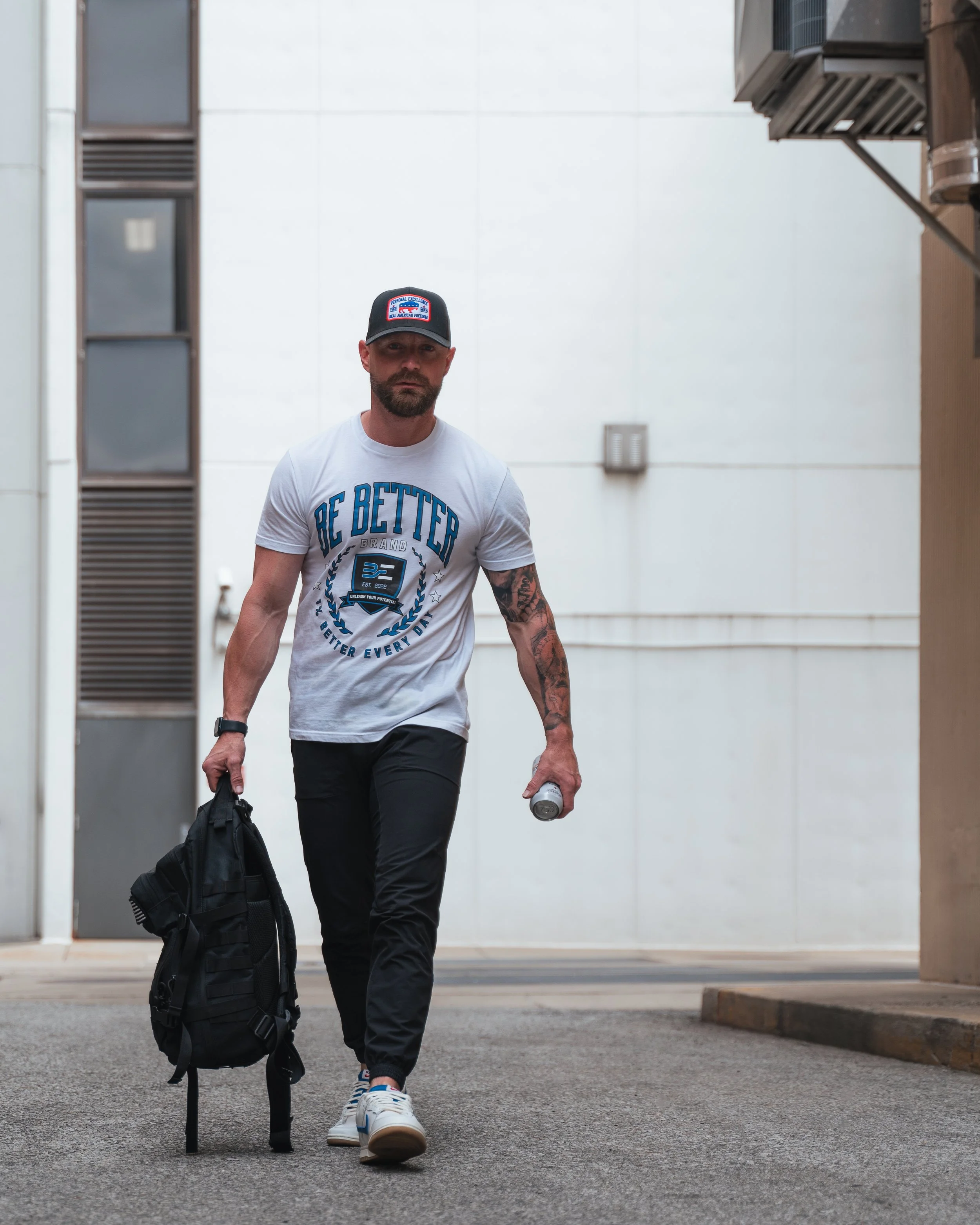 A man walking outdoors on a sidewalk, wearing a white T-shirt with blue text and graphics, a black baseball cap, black pants, and sneakers. He carries a black bag in his right hand and a water bottle in his left, with a black wristwatch on his left w