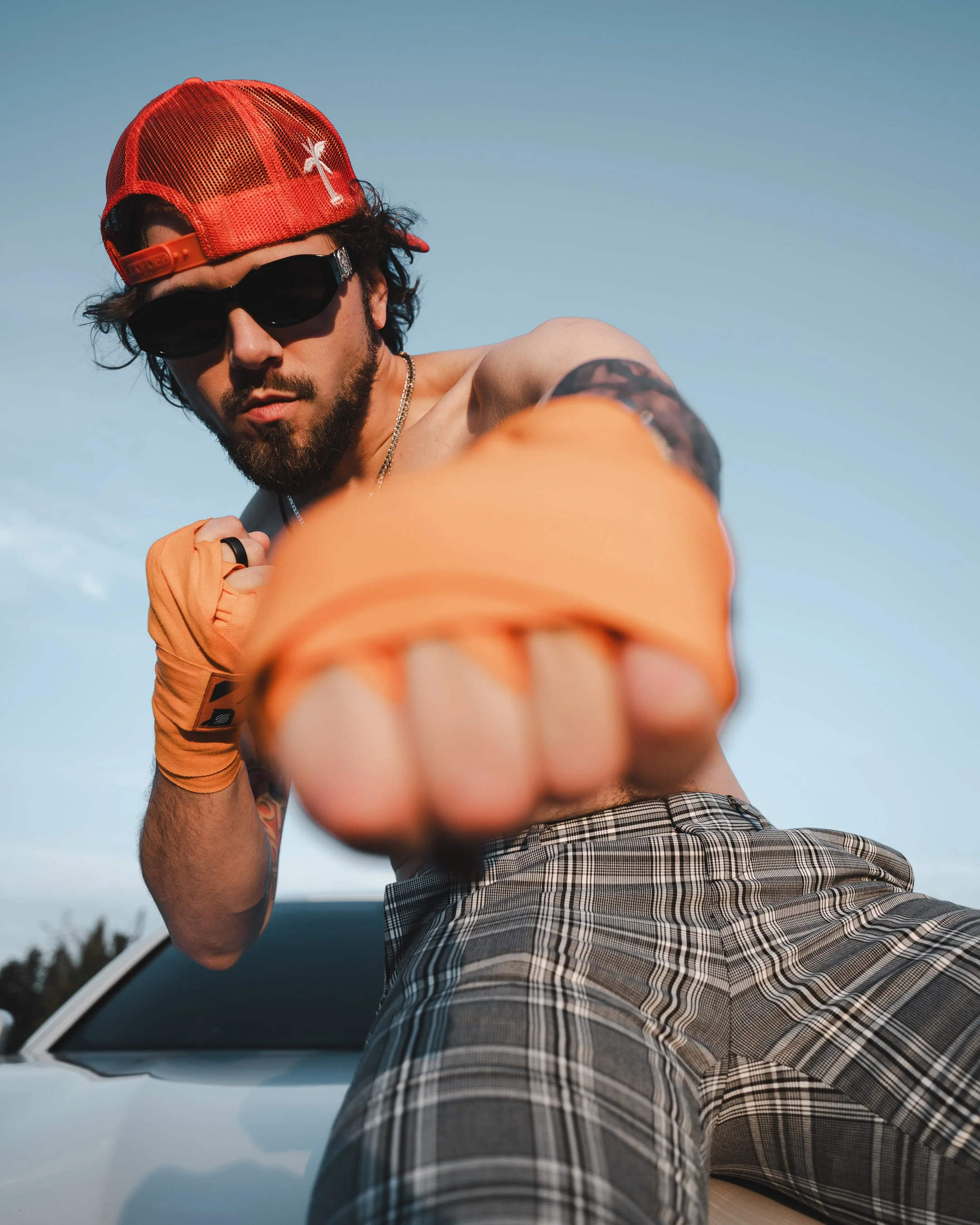 A young man with dark hair, beard, wearing sunglasses, a red cap backwards, a silver chain, and plaid shorts, is extending his arm towards the camera on a sunny day outdoors.