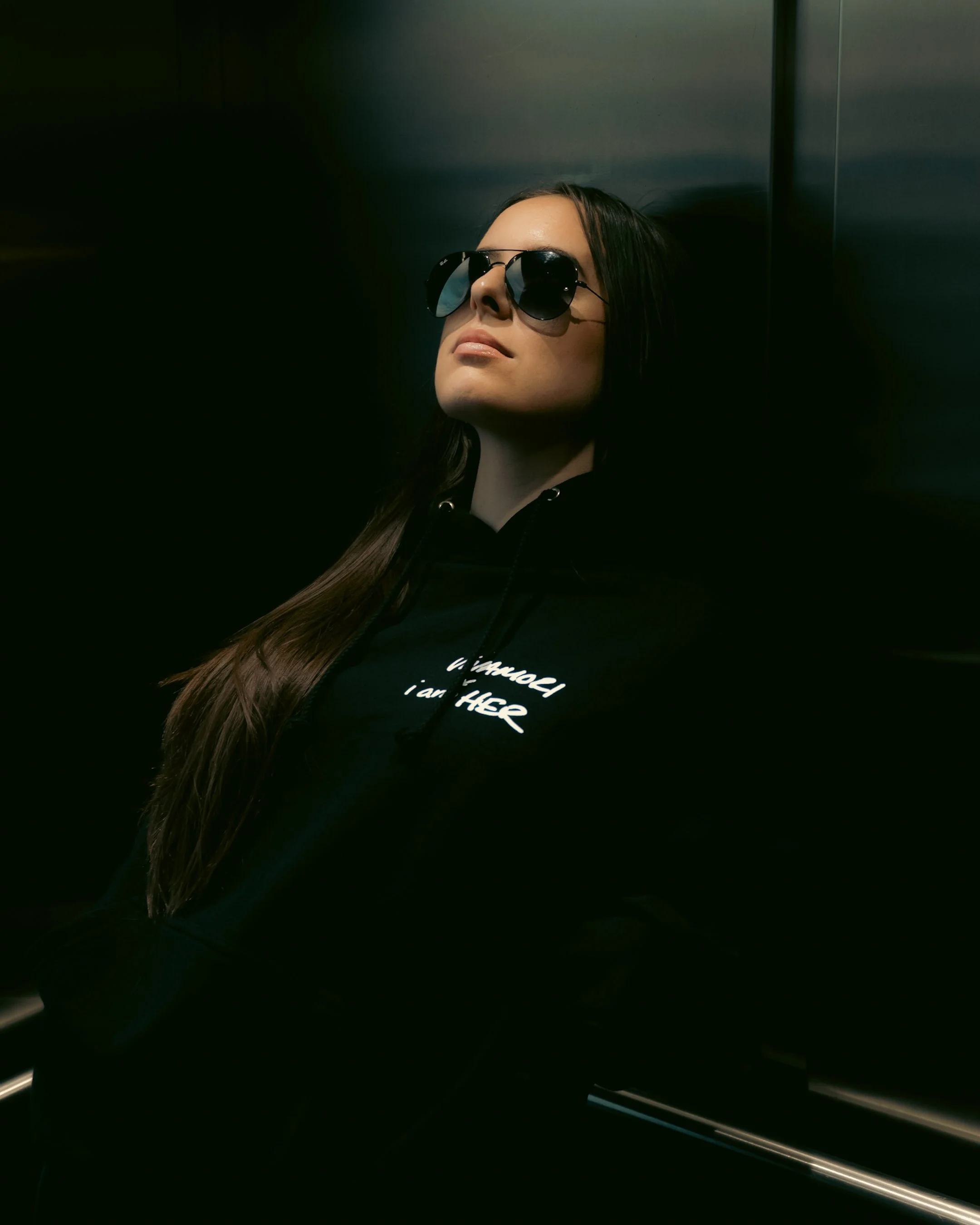 A woman with long dark hair, wearing sunglasses and a black hoodie, is in an elevator with metallic walls. She has a neutral expression and is looking upward.