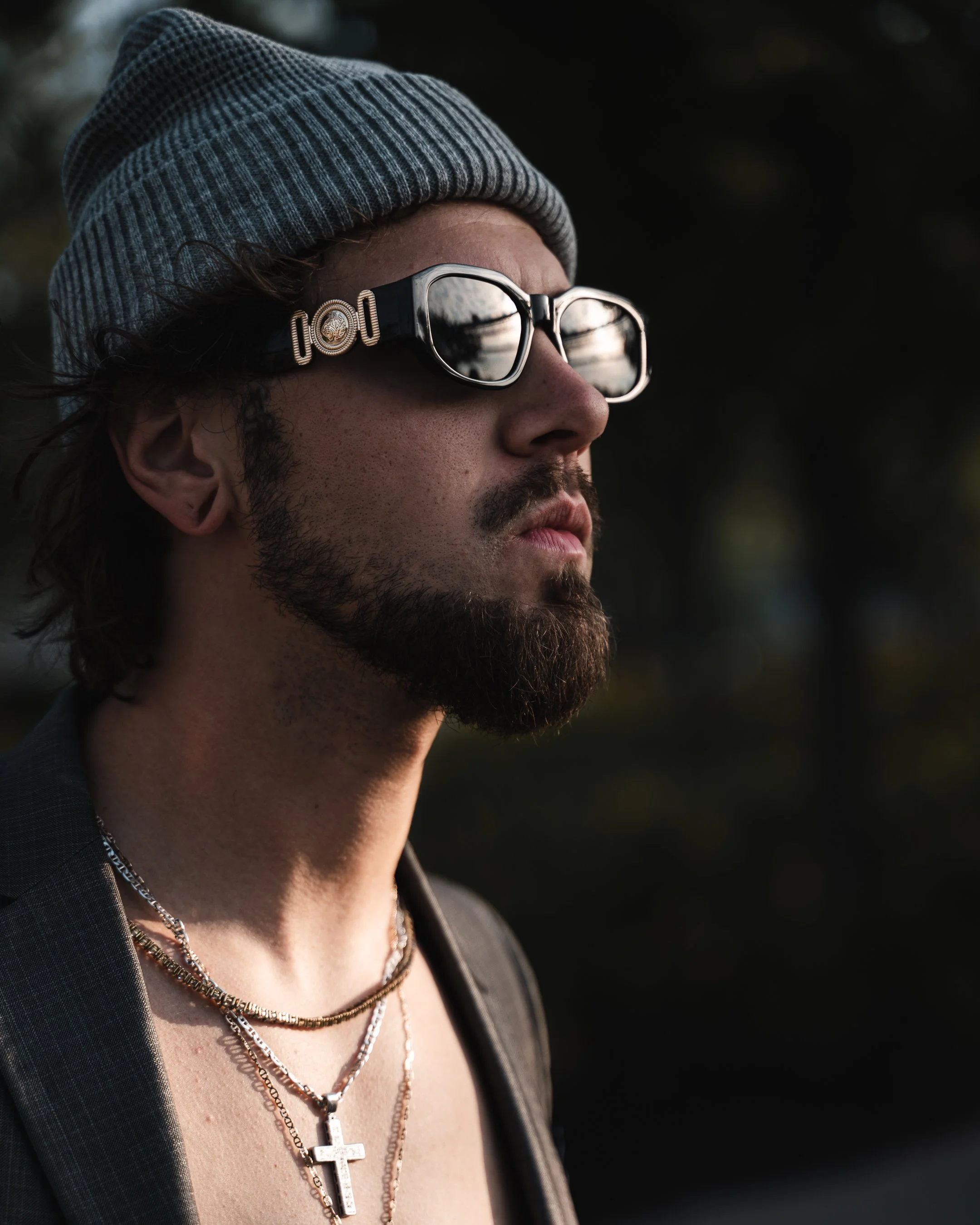 Close-up of a man wearing a gray beanie, reflective sunglasses, chains, and a cross necklace. He has a beard and mustache, and is dressed in a dark jacket with a plaid pattern.