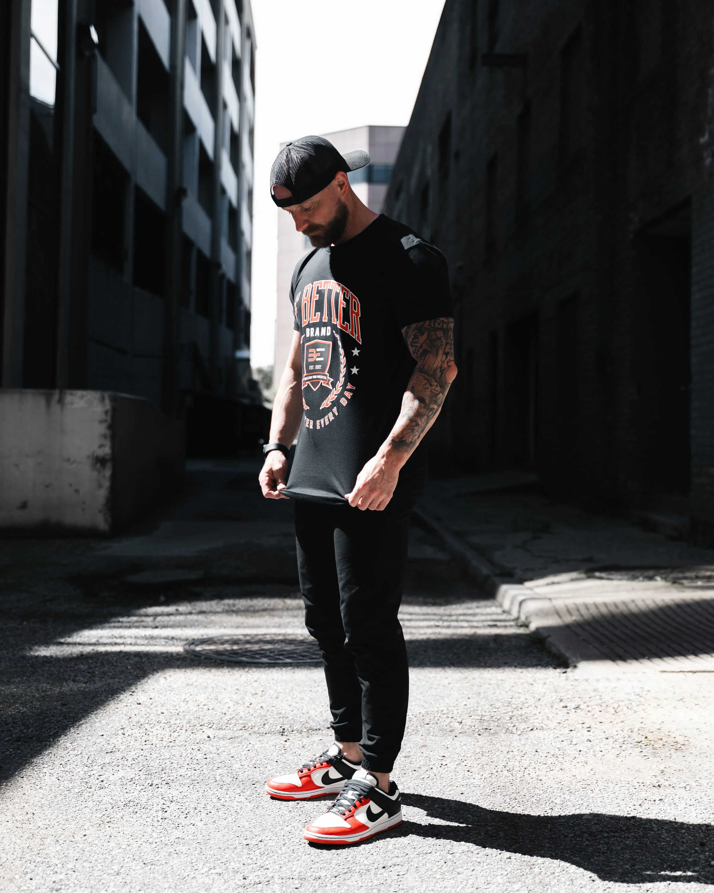 A young man with a beard and tattoos wearing a black cap, black T-shirt, black pants, and red, white, and black sneakers, standing outdoors on a paved sidewalk between dark buildings, looking down and adjusting his shirt.