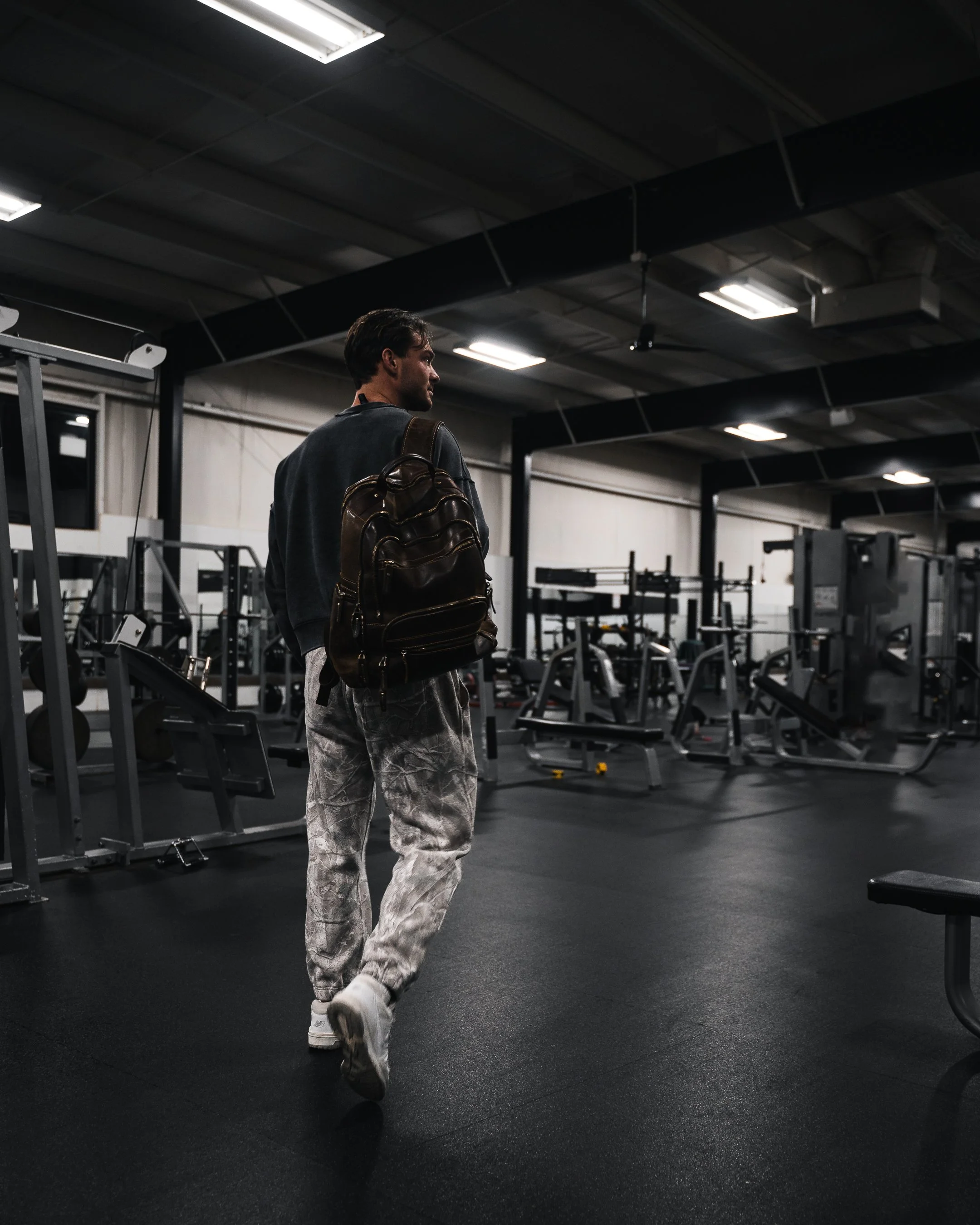 A young man in camouflage pants, white sneakers, and a dark jacket with a brown backpack walking inside a gym. The gym has various exercise equipment, including weight machines and racks, with a dark ceiling and fluorescent lighting.