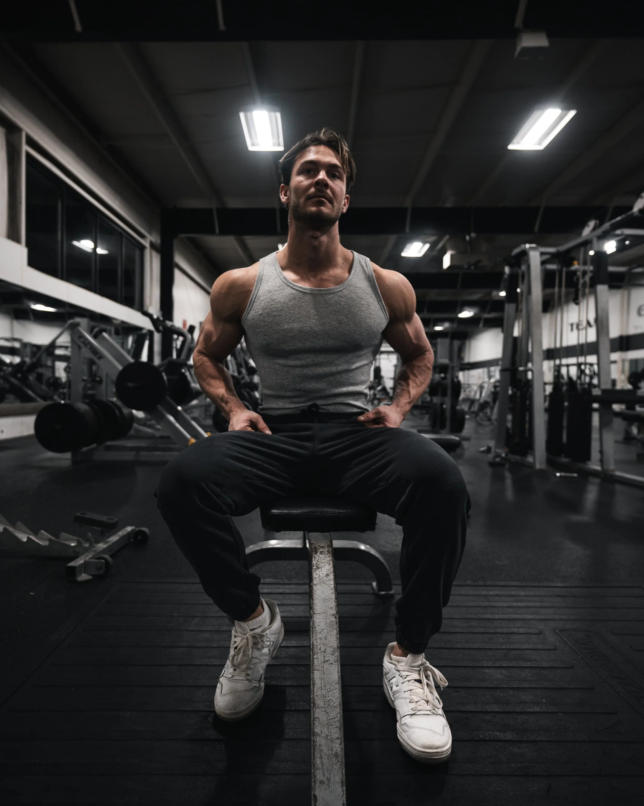 A muscular man in a gray tank top and black pants sitting on a workout bench in a gym, with weightlifting equipment around him and low ceiling lights overhead.