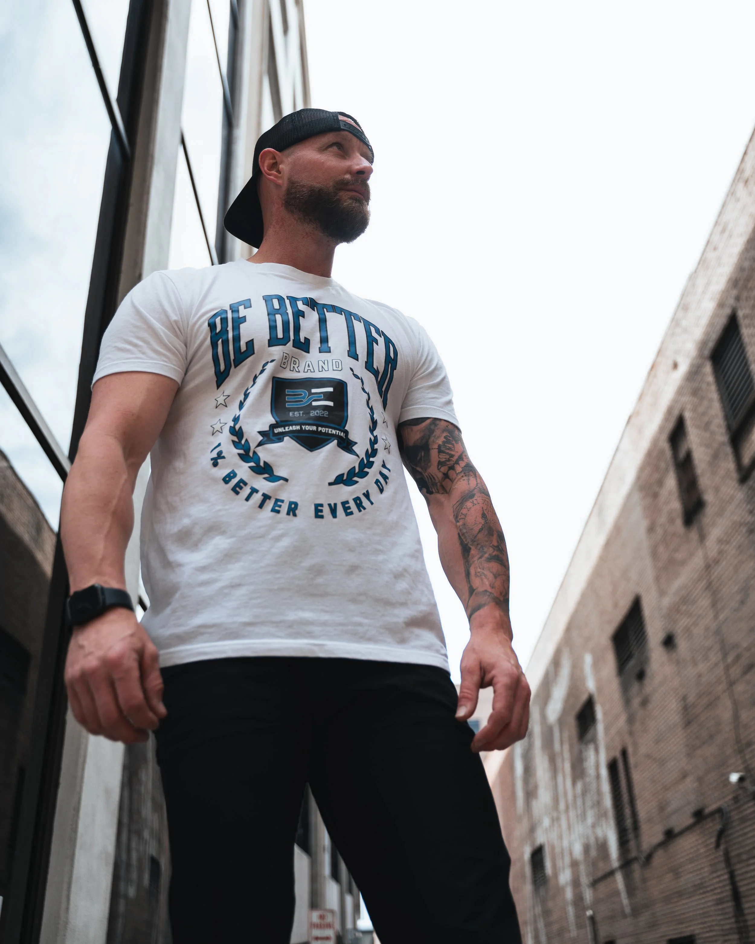 A man with a beard wearing a black baseball cap, white T-shirt with blue lettering, and tattoos on his arm, standing in an alley between brick buildings, looking upward.