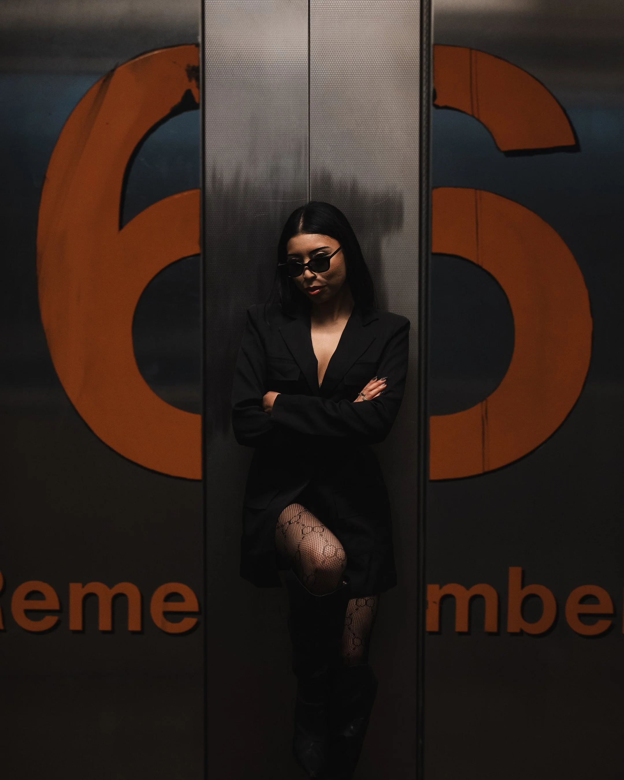A woman with black hair, wearing sunglasses, a black blazer, fishnet stockings, and black boots, standing with her arms crossed in an elevator with a reflective metallic interior and an orange logo on the doors.