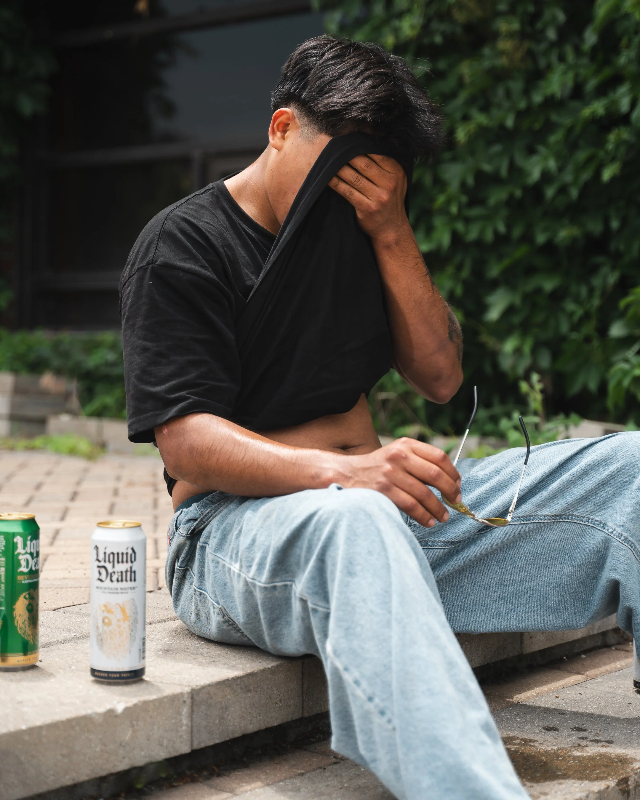 A young man sitting on a brick ledge with three cans of Liquid Death sparkling water in front of him, covering his face with his shirt, outdoors with greenery in the background.