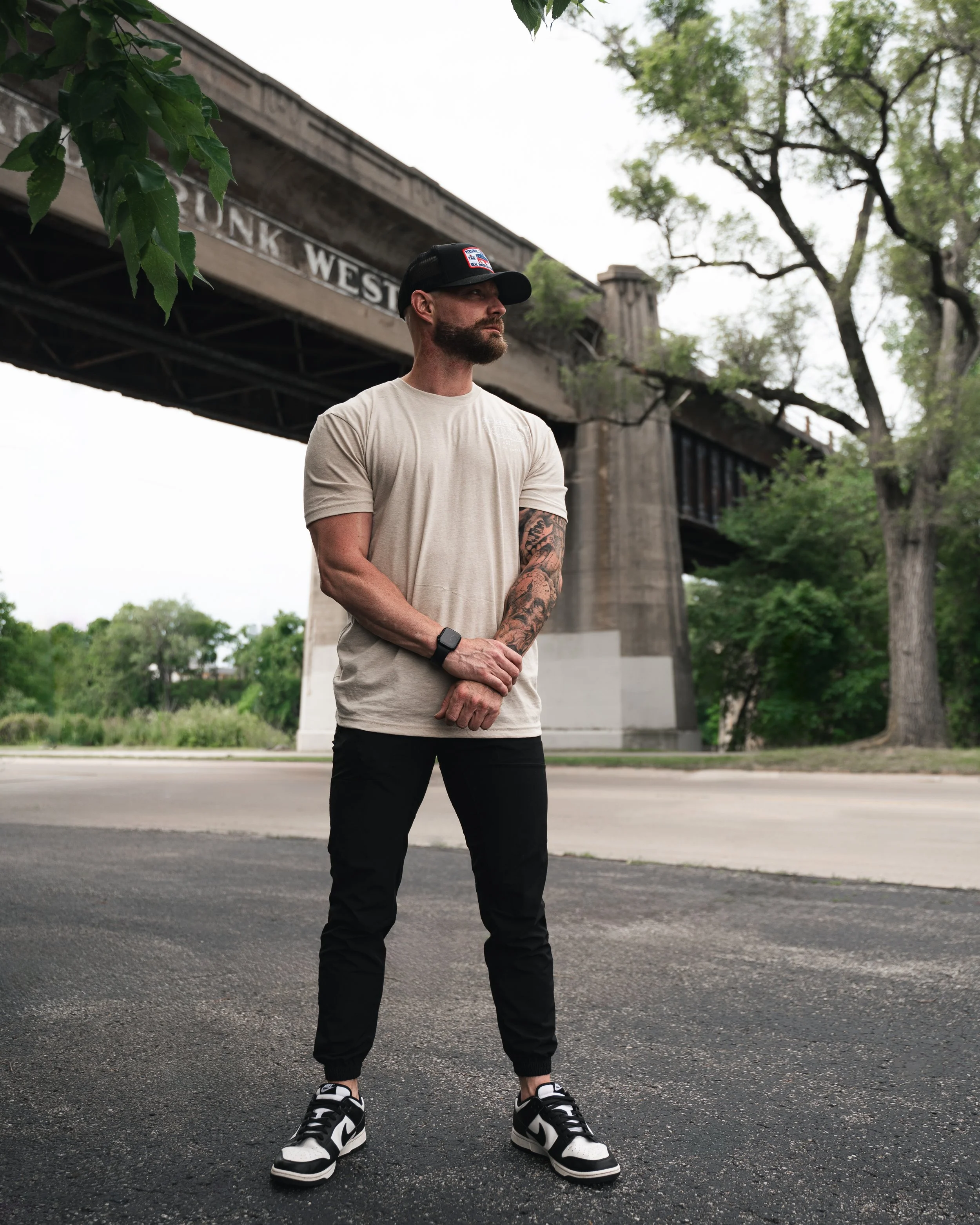 Man with a beard and tattoos wearing a beige t-shirt, black pants, and sneakers standing outdoors under a bridge with trees in the background.