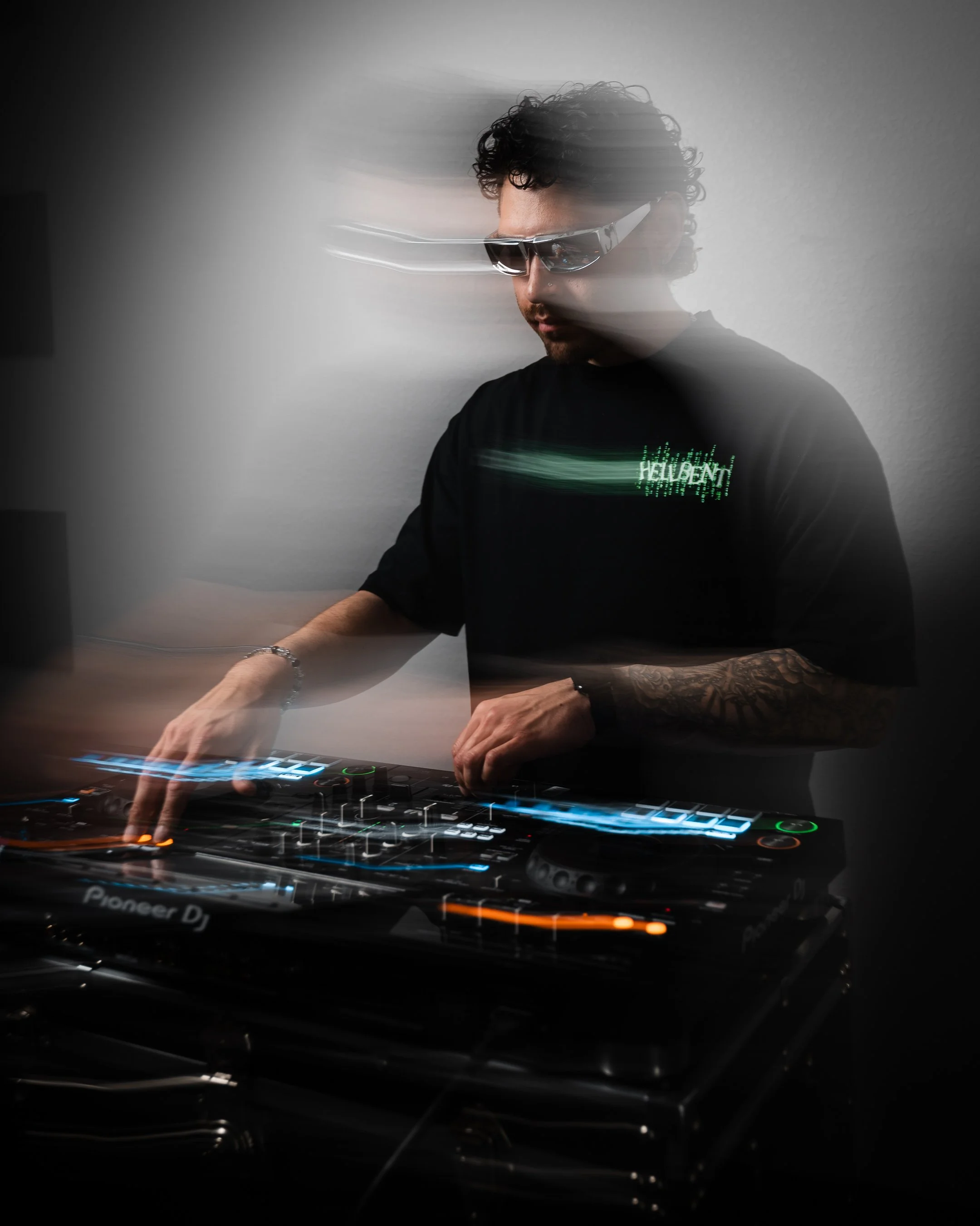 A DJ wearing sunglasses and a black shirt with green digital text is operating a Pioneer DJ controller in a dark setting, with motion blur indicating movement.