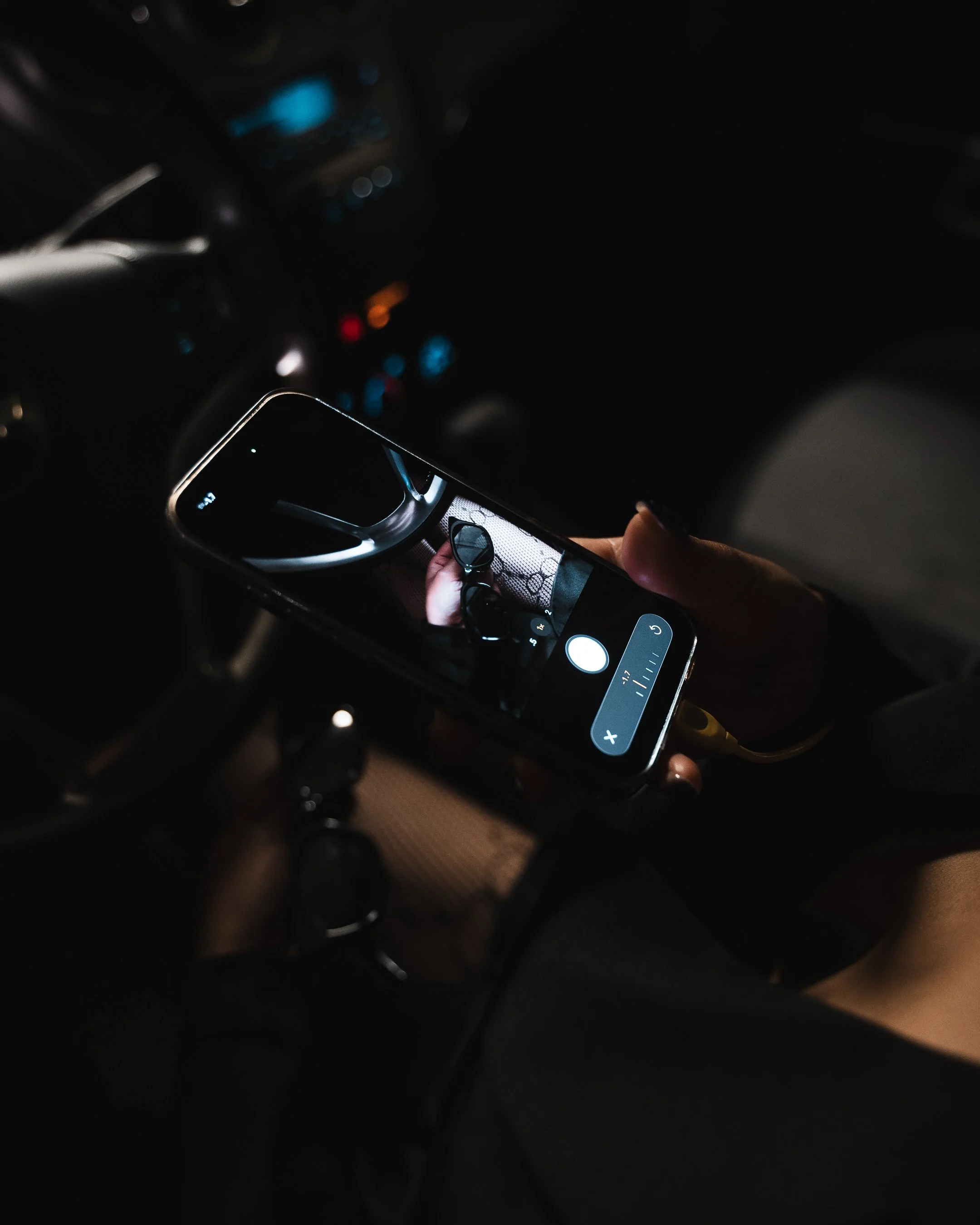 A person holding a smartphone in a dimly lit car interior. The phone screen shows an image of someone, but the background and details are obscured by darkness.