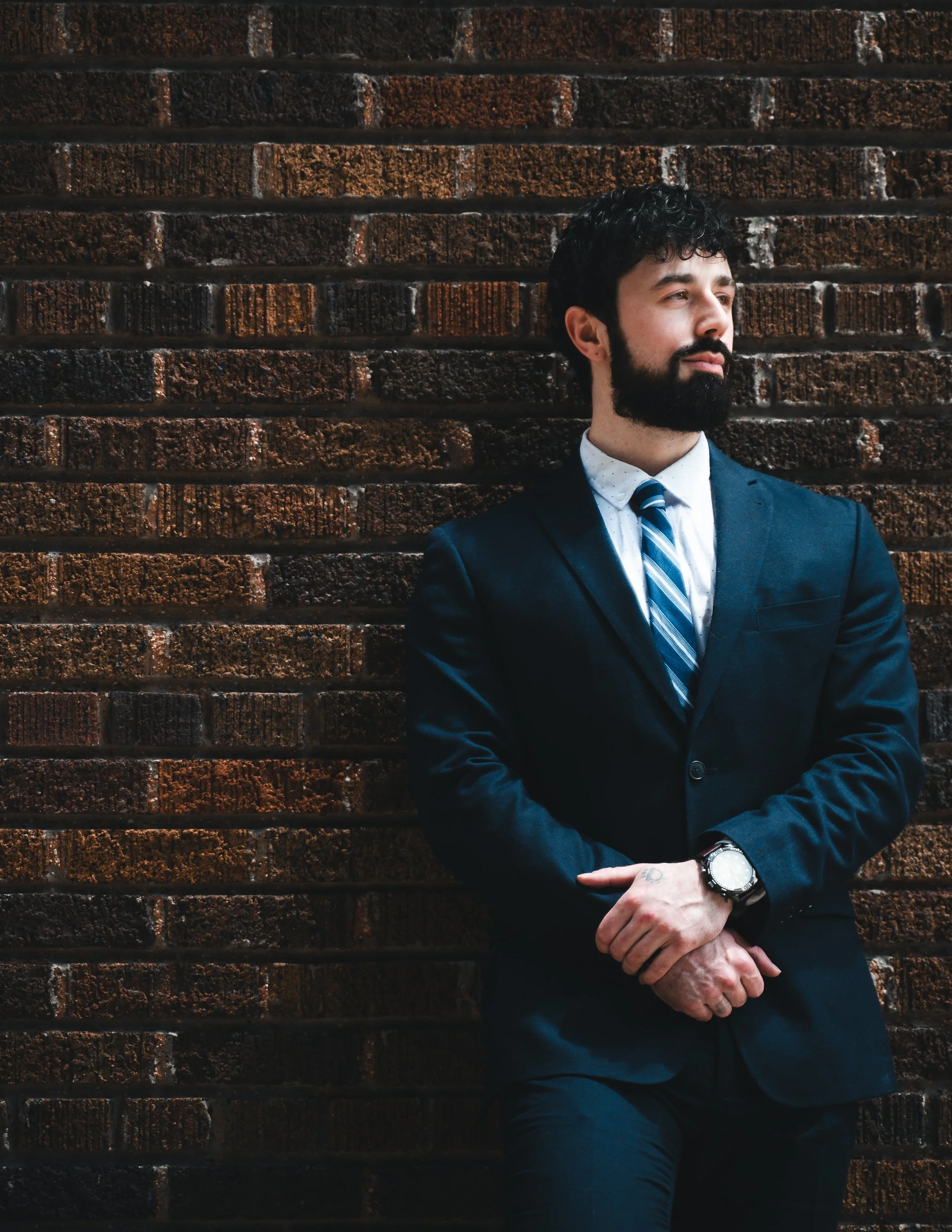 A man in a navy suit with a white shirt and striped tie is standing against a red brick wall, with his arms crossed and looking to his right. He has dark curly hair, a beard, and is wearing a wristwatch.