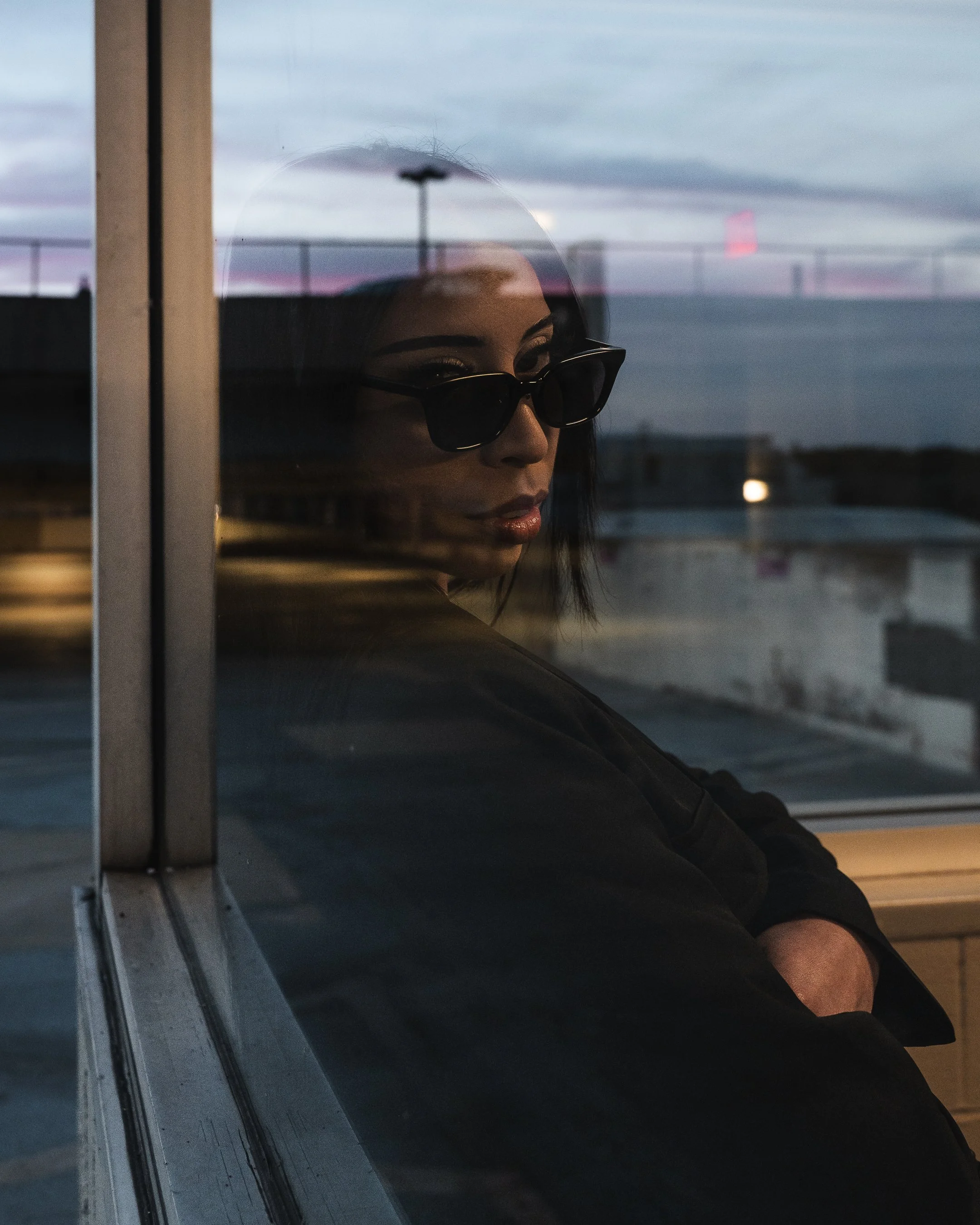 Woman wearing sunglasses looking out of a window on a cloudy evening, with a cityscape and a parking lot visible outside.