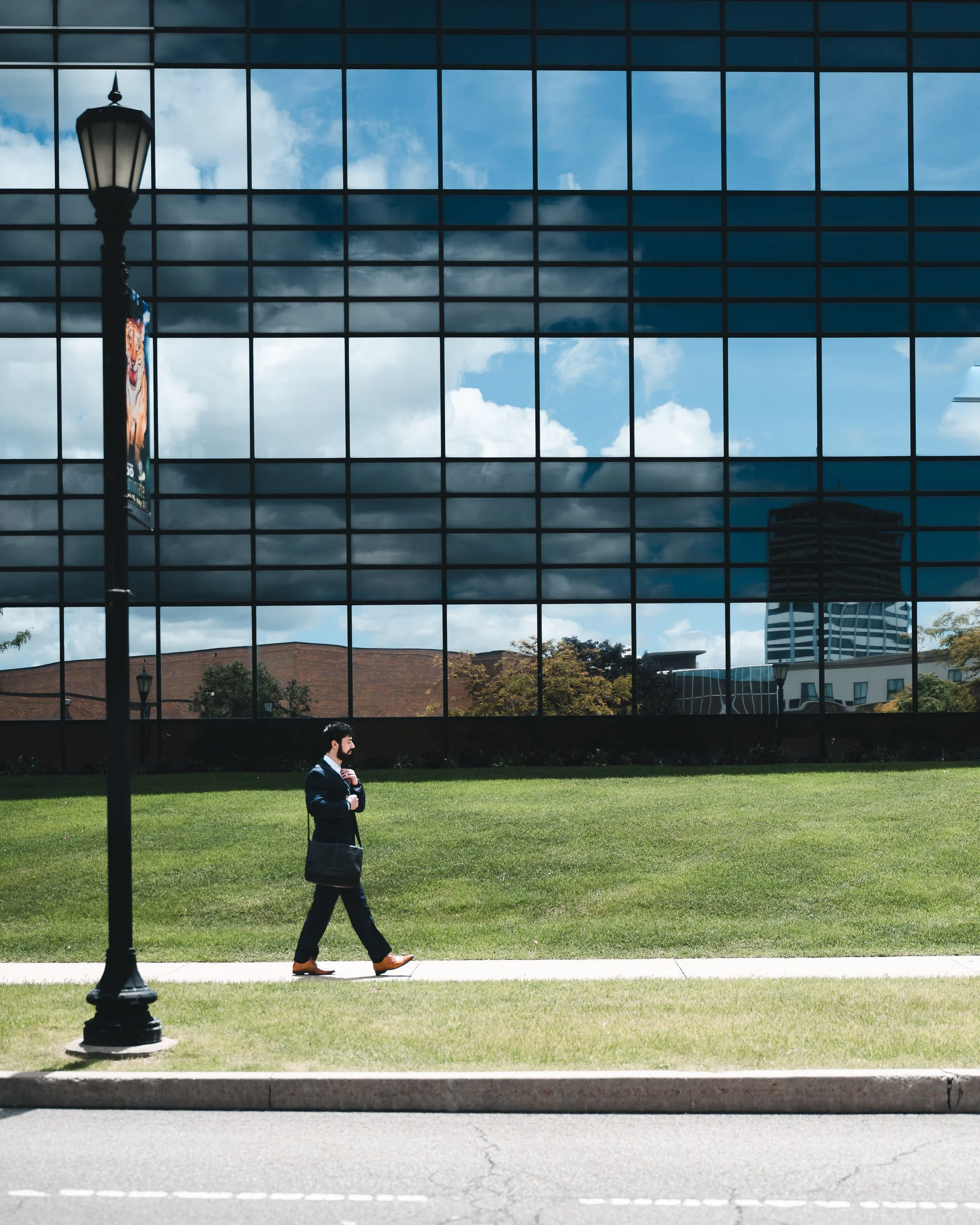 A man in a suit walking on a sidewalk with a briefcase in front of a large glass building reflecting the sky and clouds, a street lamp with an advertisement, and trees and buildings in the background.