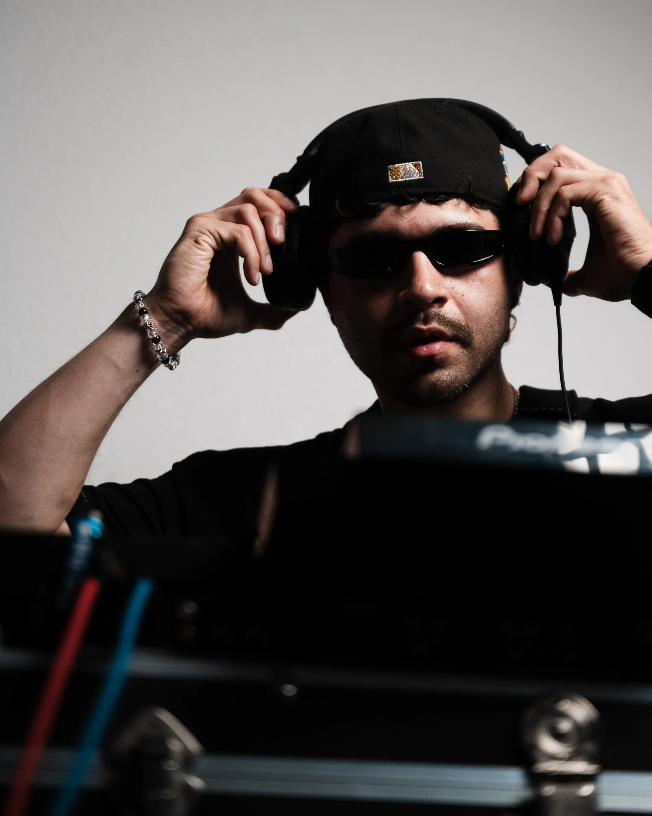 Man wearing sunglasses and headphones, adjusting headphones over his cap, with audio equipment in front.