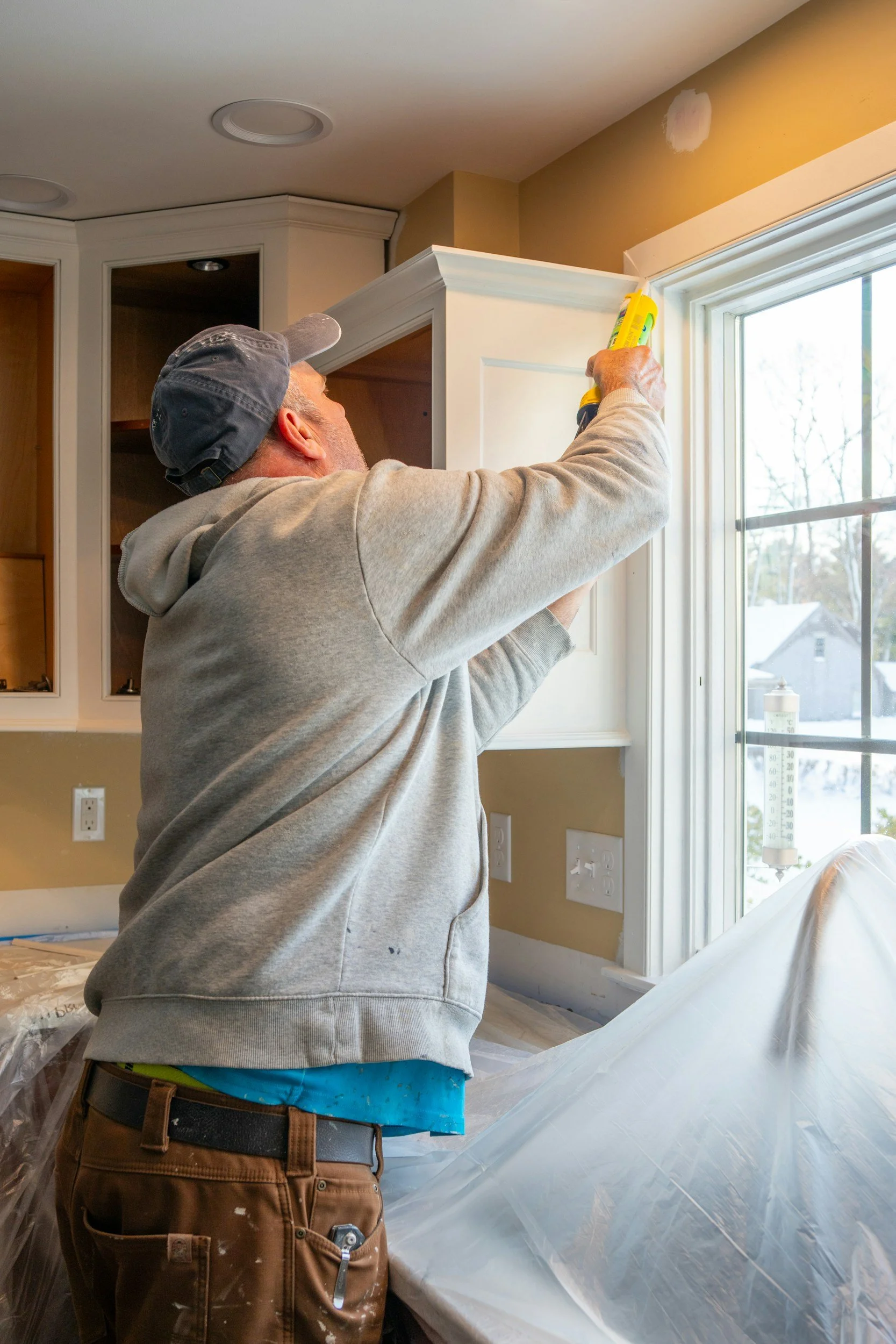 A man wearing a gray hoodie, brown work pants, and a cap is using a caulking gun to apply white sealant or paint along the top edge of a kitchen cabinet next to a window.