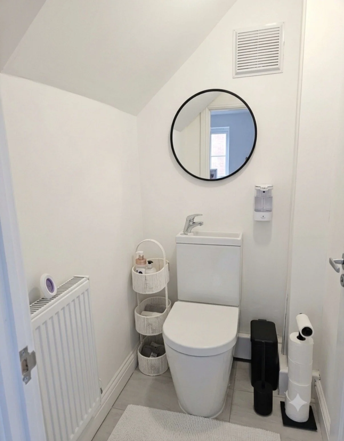 Small bathroom with white walls, a round mirror, a compact sink, and a toilet. There are small storage baskets on a tiered stand, a soap dispenser, a toilet paper holder, a radiator, and various bathroom accessories.