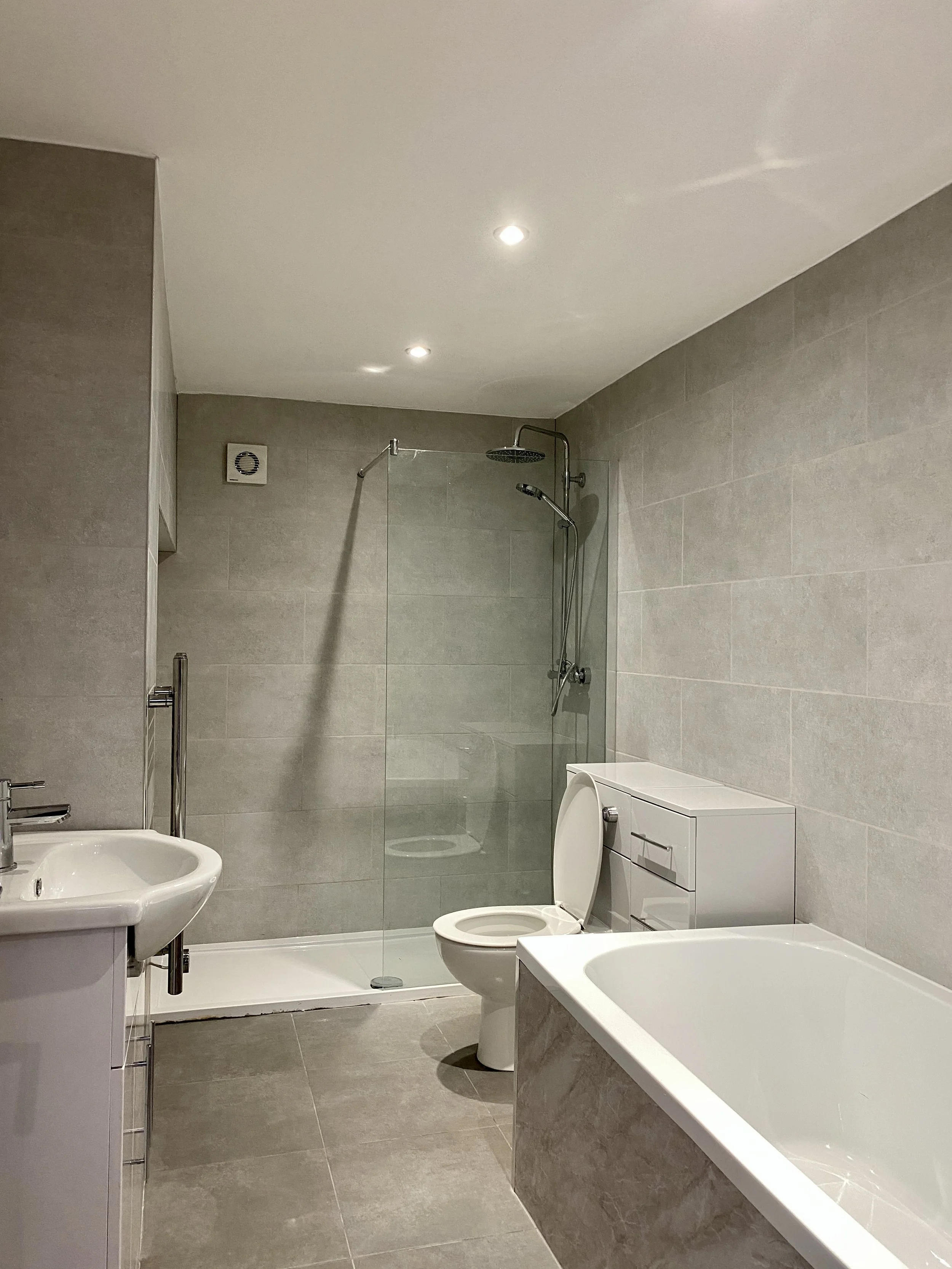 A modern, minimalist bathroom with a bathtub, toilet, small bathroom sink, and a walk-in shower with glass door, gray tiled walls, and ceiling lights.