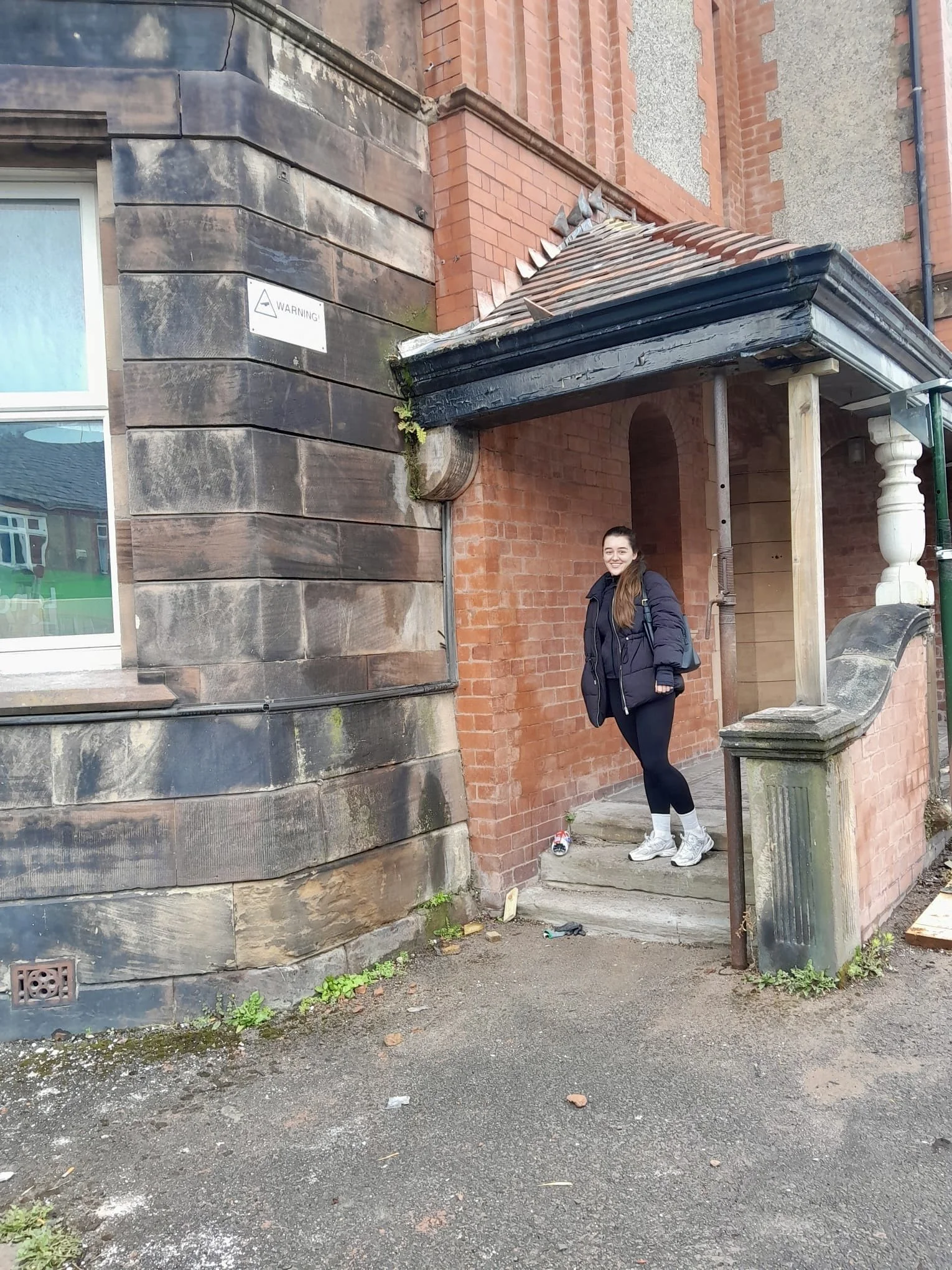 A young woman with long brown hair wearing a dark winter coat, black leggings, and white sneakers stands on a stone step outside a brick building. She is smiling and looks happy.