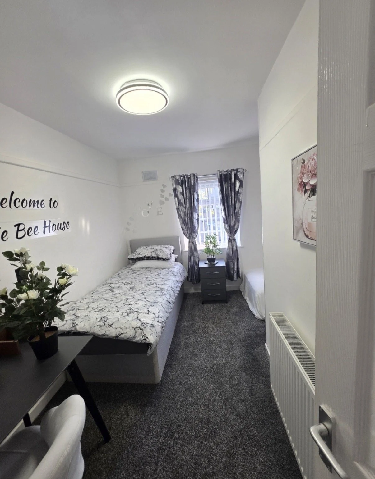 A cozy guest bedroom with a single bed, black and white bedding, a small black nightstand with a plant, and a window with black and white patterned curtains, decorated with wall art and flowers.
