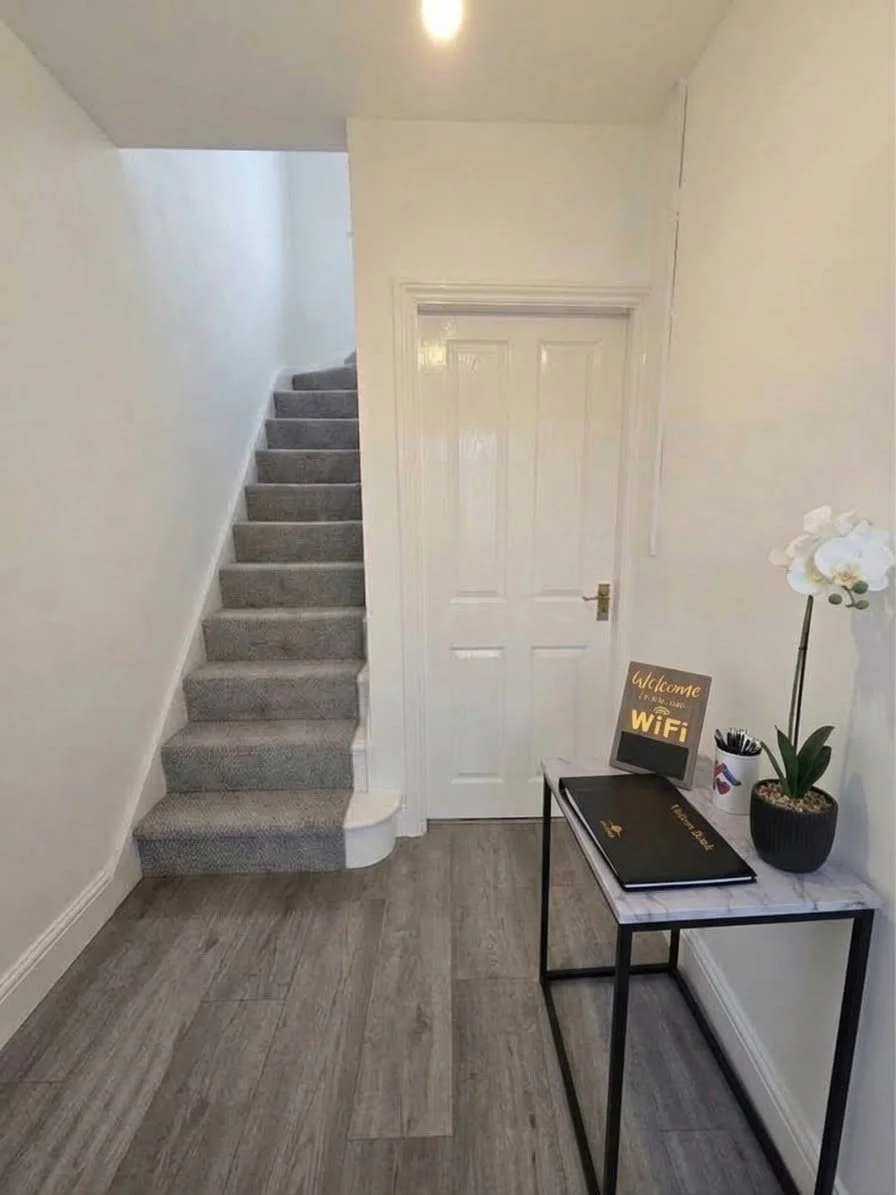 Entryway with a staircase to the left, closed door in the center, small table with a welcome WiFi sign, potted orchid, container with pens, and notebooks on the right