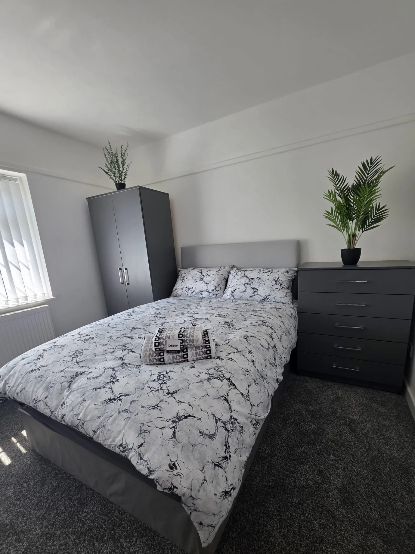 A neatly made bed with a marble-patterned duvet cover, flanked by two dark gray dressers with potted plants on top, in a minimalist bedroom with a window on the left and white walls.