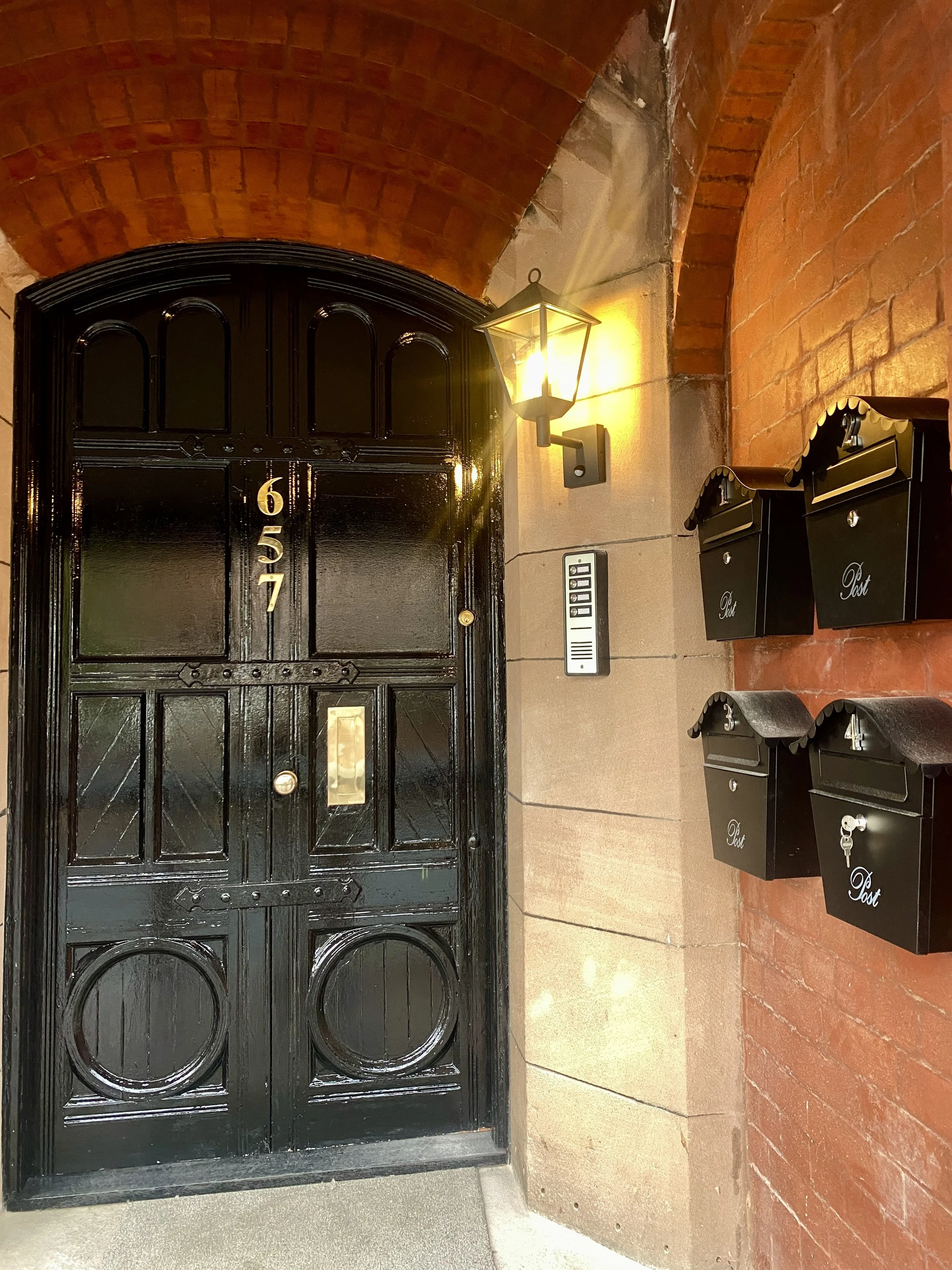 Black front door with the number 657 on it, mounted on a stone wall. To the right are black mailboxes labeled 'Post', mounted on a brick wall. There is an outdoor lantern light illuminating the entrance, and an intercom system is also visible.