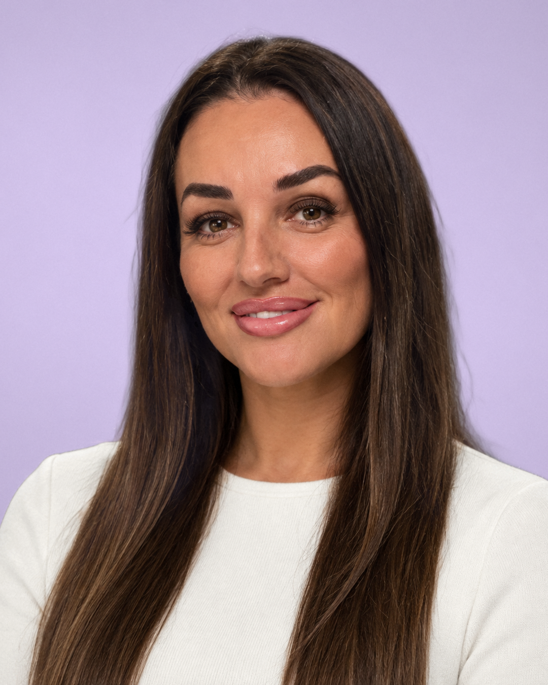 A woman with long brown hair and makeup, smiling in front of a light purple background.